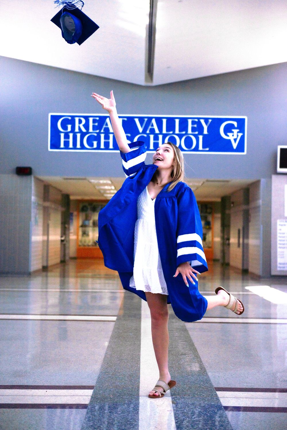 Person in blue graduation gown dances joyfully in high school hallway during graduation photoshoot.