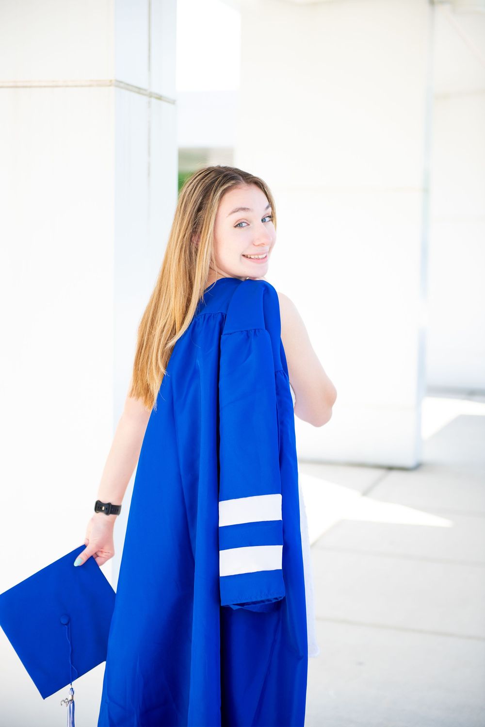 Graduate in bright royal blue academic regalia holding cap while looking back over shoulder in bright setting.