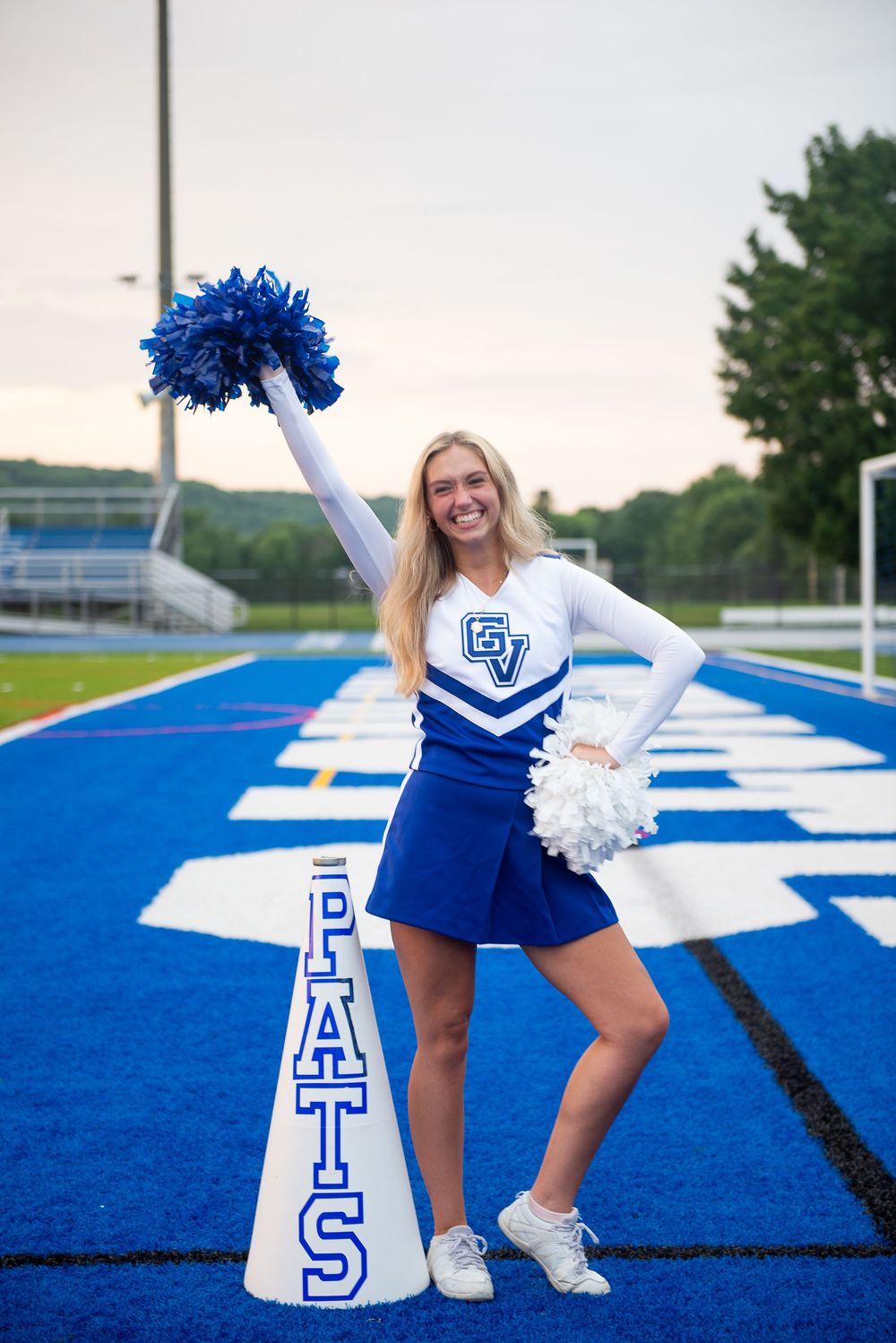 A cheerleader in blue and white uniform poses with pom poms and a megaphone prop on a blue football field.
