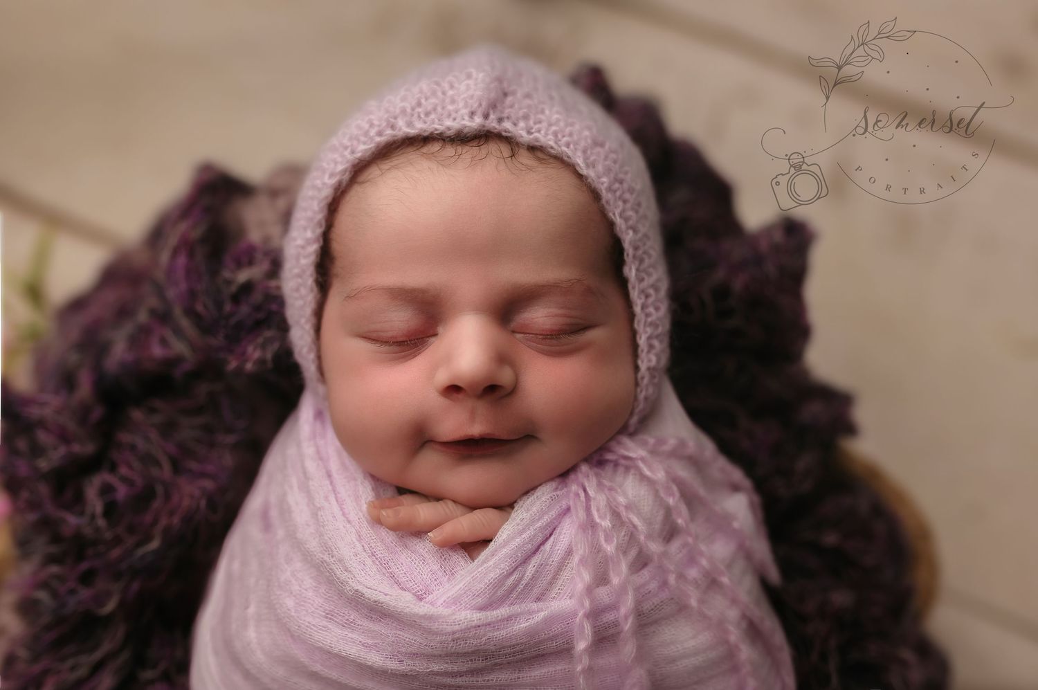 Newborn baby girl wrapped in purple with matching hat, smiling softly at Somerset Portraits studio in Charlottesville, VA.
