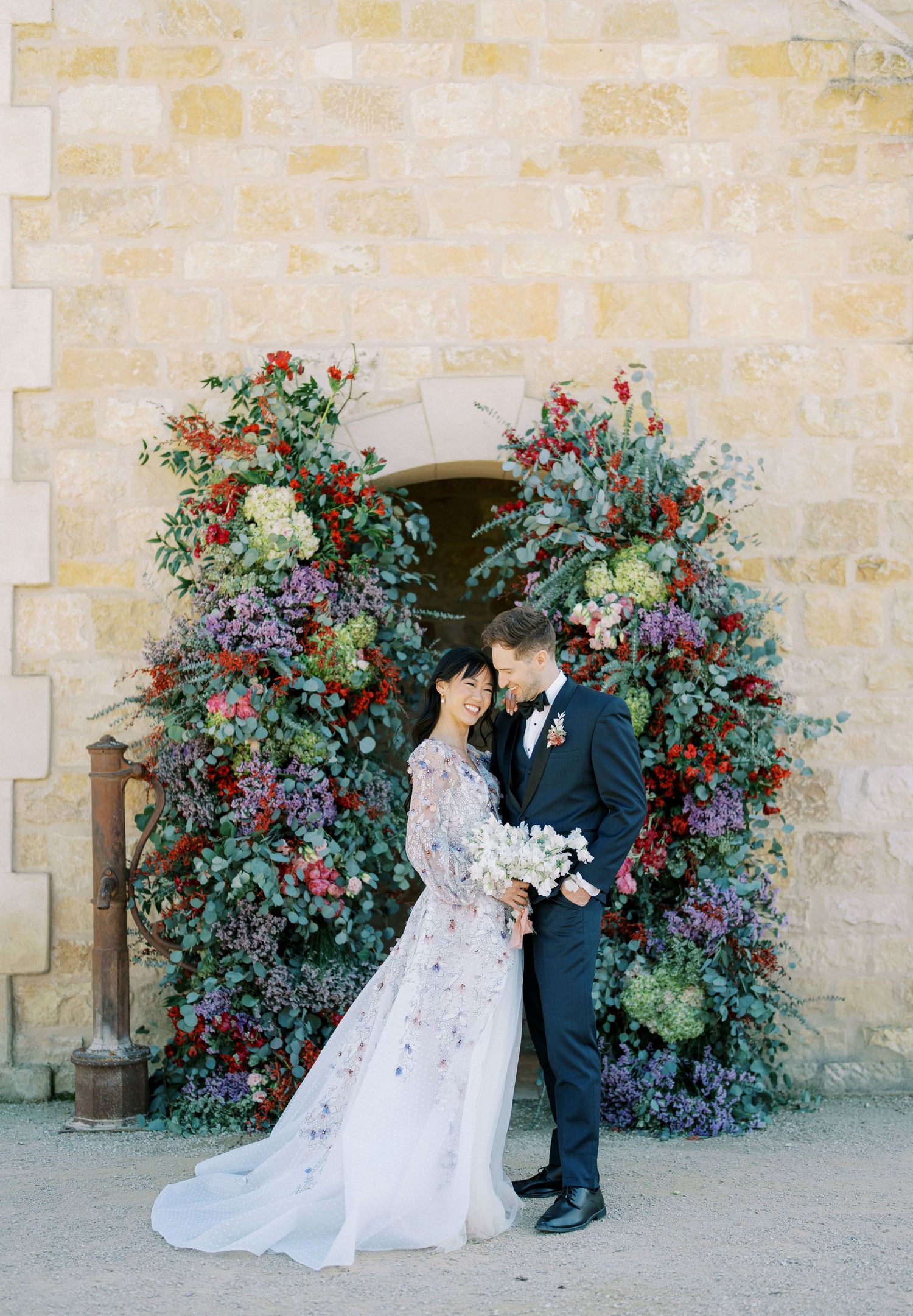 A wedding portrait under a stunning floral arch with blue and red blooms against stone walls.