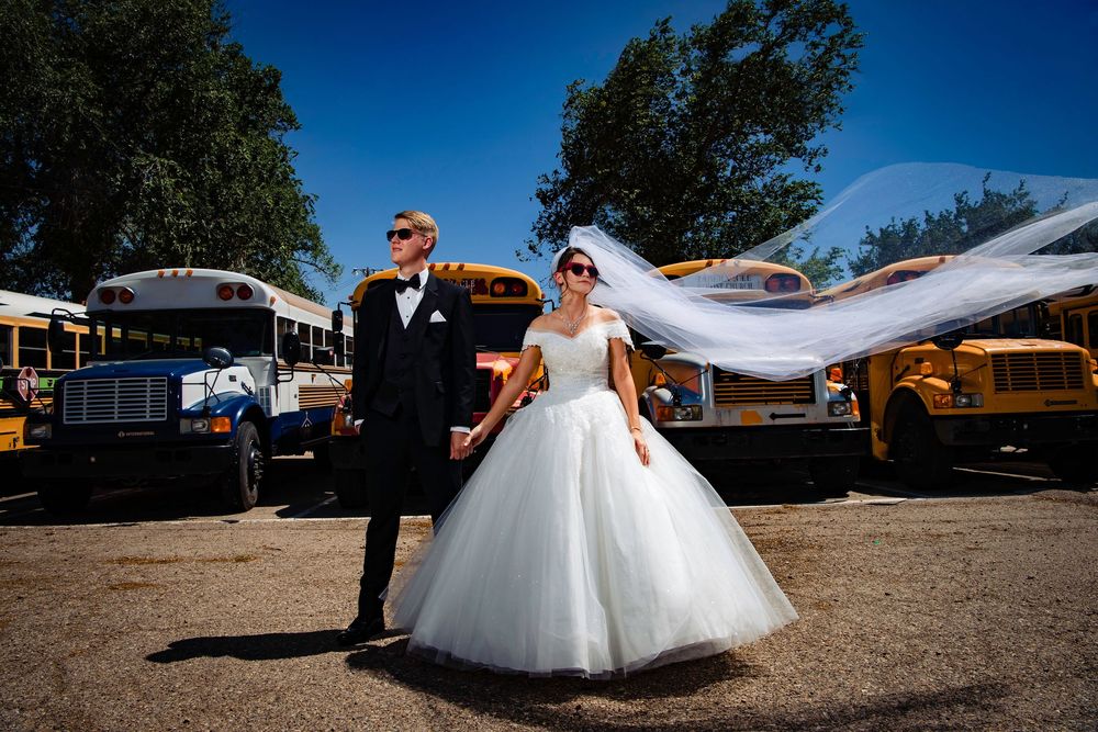 Wedding couple poses in panoramic outdoor setting with flowing white wedding dress against blue sky and rustic backdrop.
