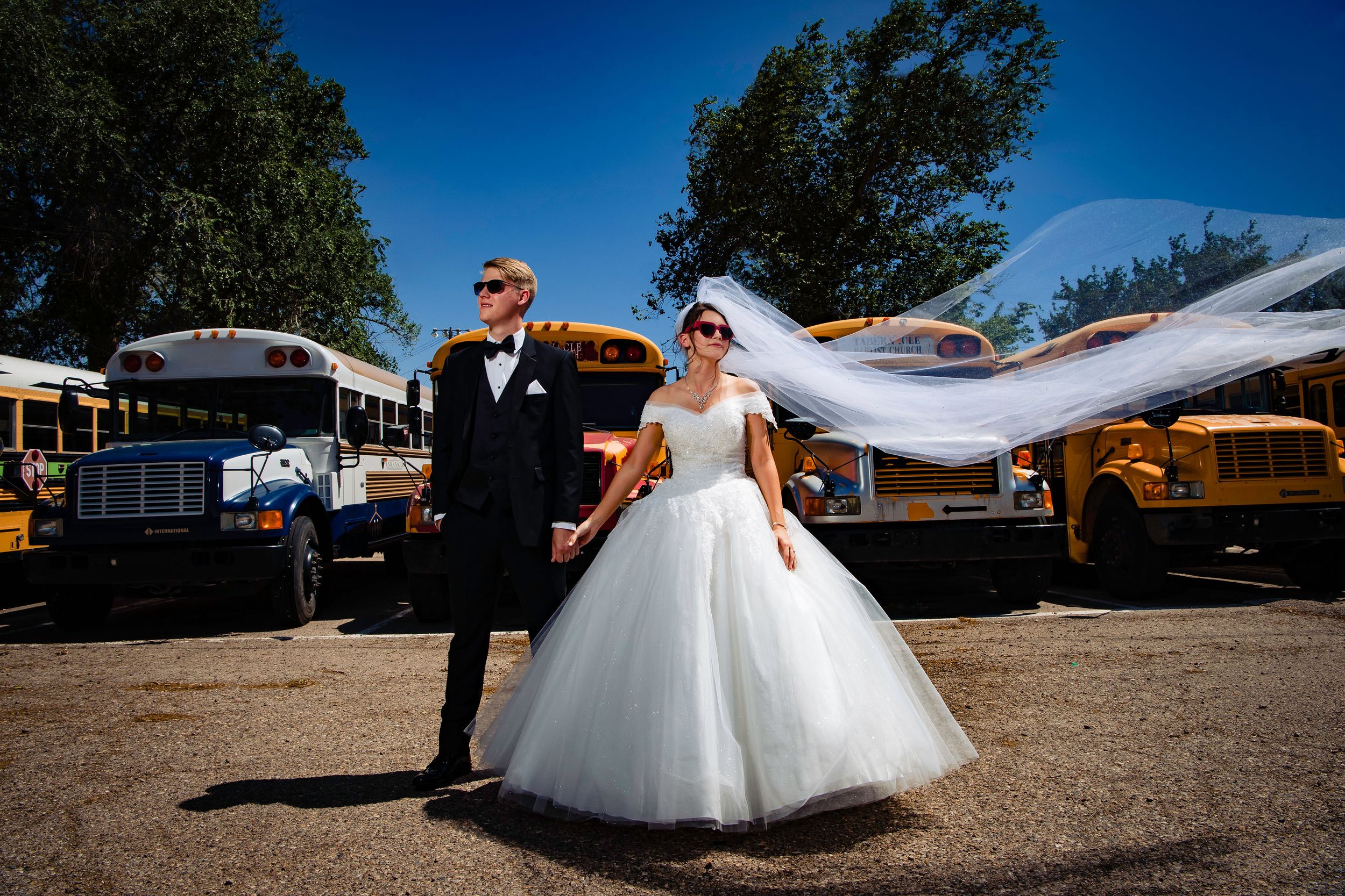 Wedding couple poses in panoramic outdoor setting with flowing white wedding dress against blue sky and rustic backdrop.