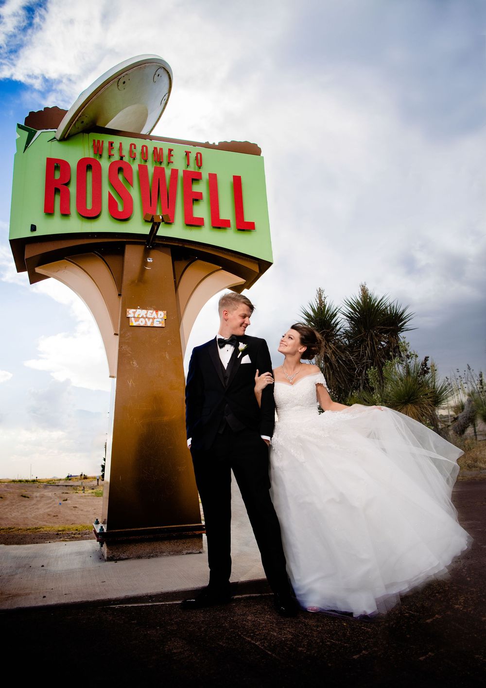 A couple in wedding attire poses under the iconic 'Welcome to Roswell' sign against a cloudy New Mexico sky.