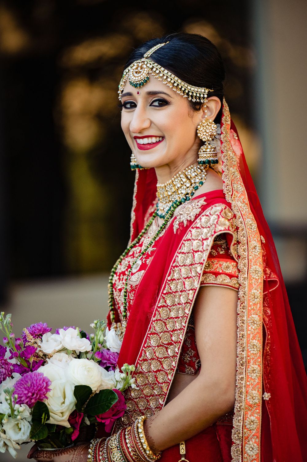 Traditional Indian bride in red lehenga wearing ornate gold jewelry and headpiece holds a colorful floral bouquet.