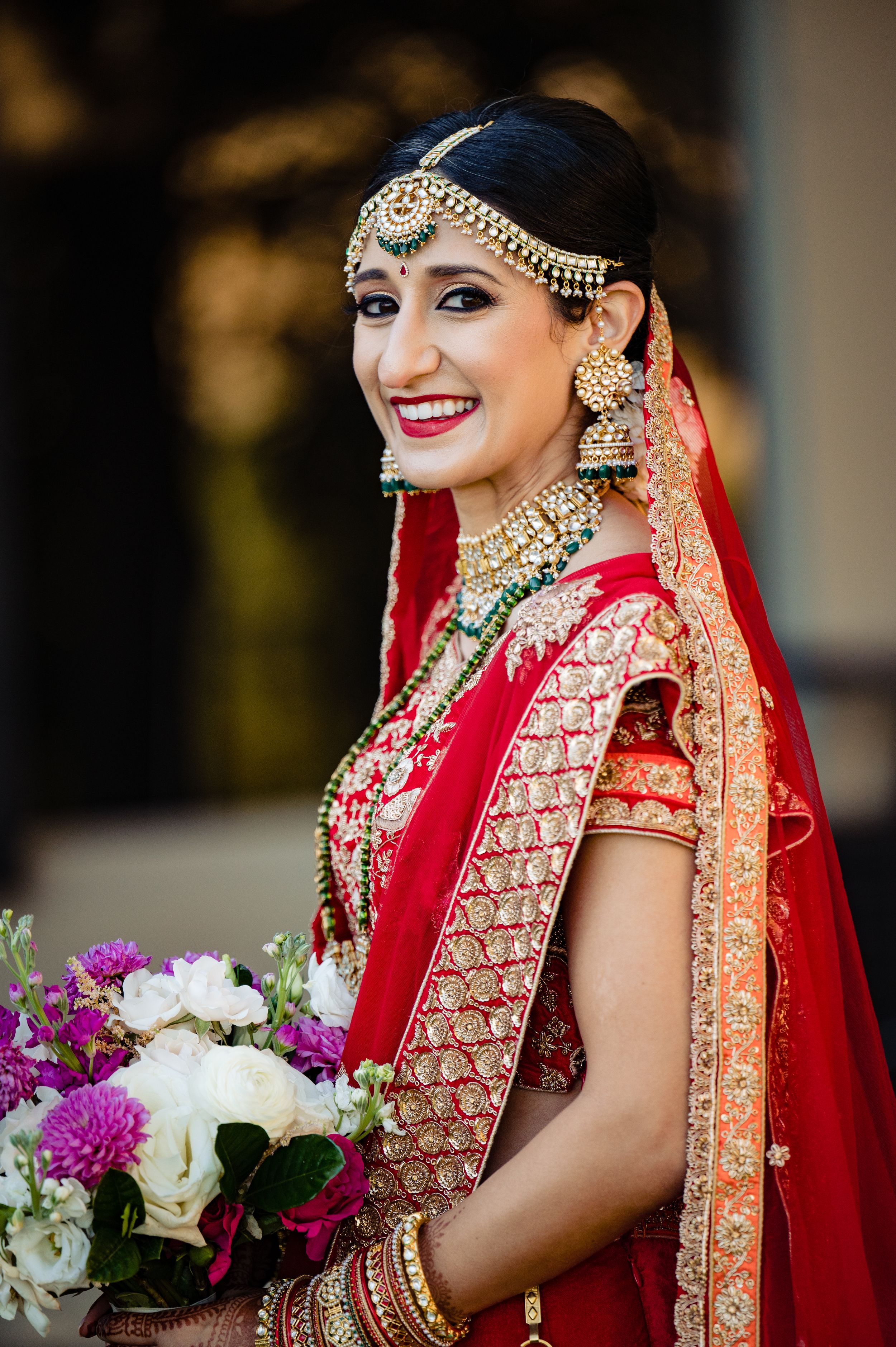 Traditional Indian bride in red lehenga wearing ornate gold jewelry and headpiece holds a colorful floral bouquet.