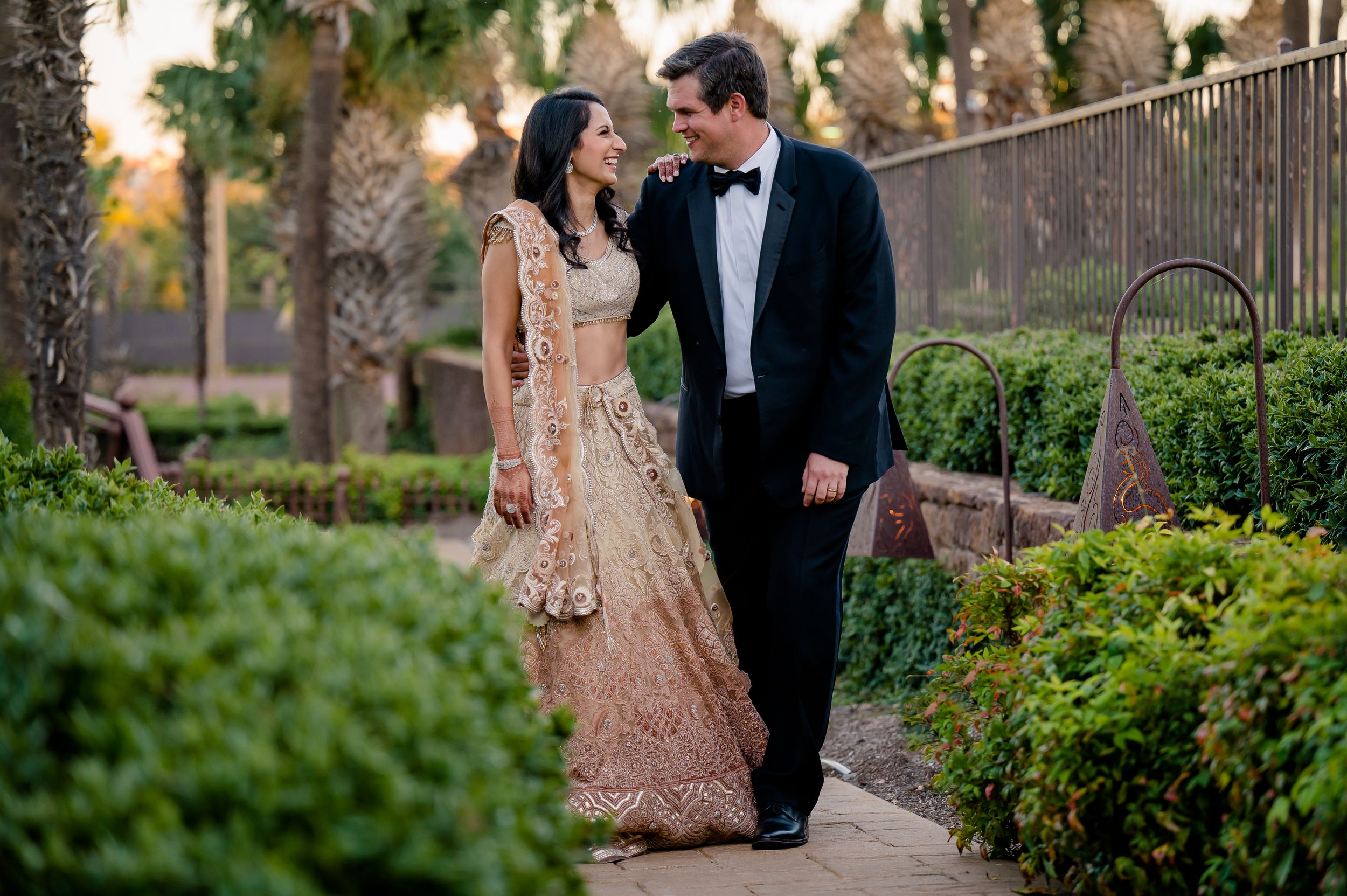 A couple in formal attire, with the woman wearing an ornate peach lehenga, pose together on a garden path at sunset.