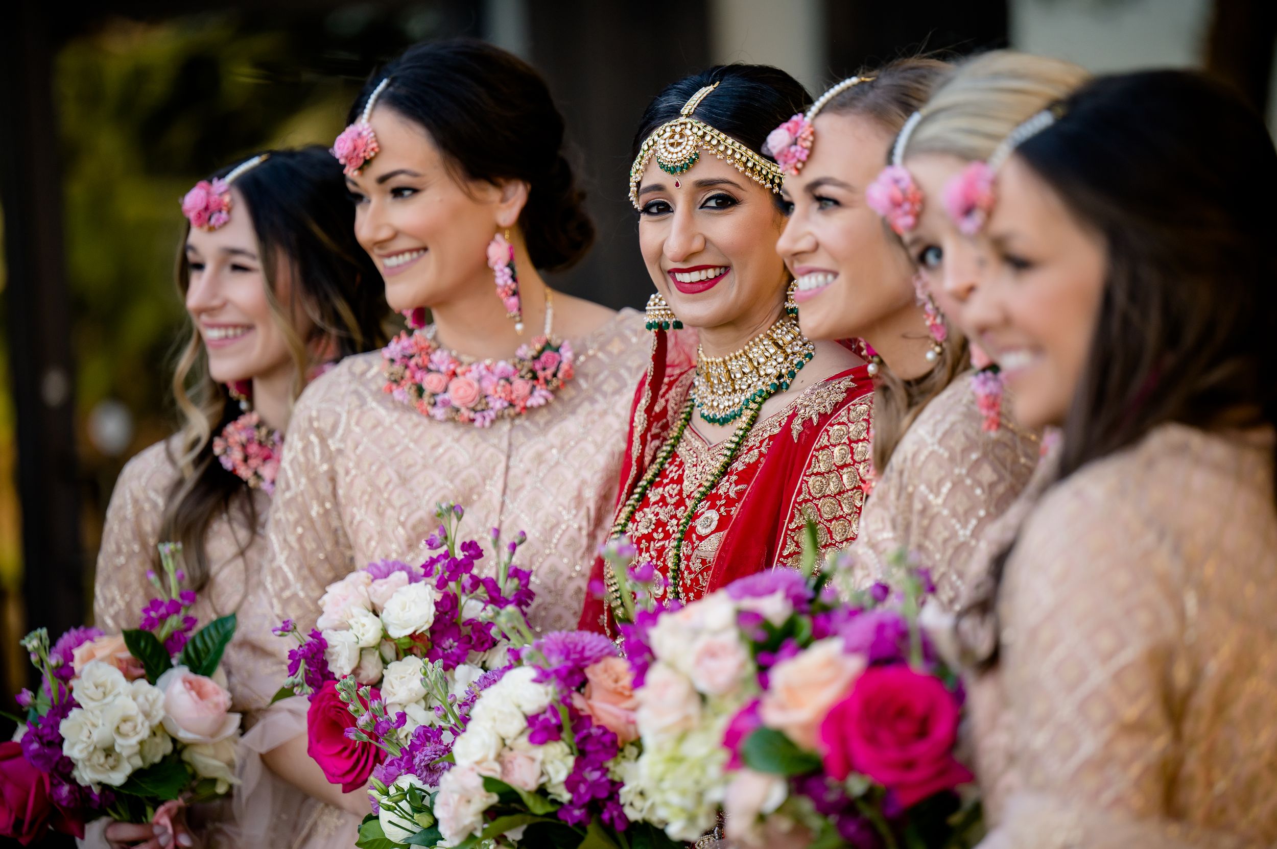 A bridal party in South Asian wedding attire holds vibrant pink and white floral bouquets while sharing a joyful moment together.