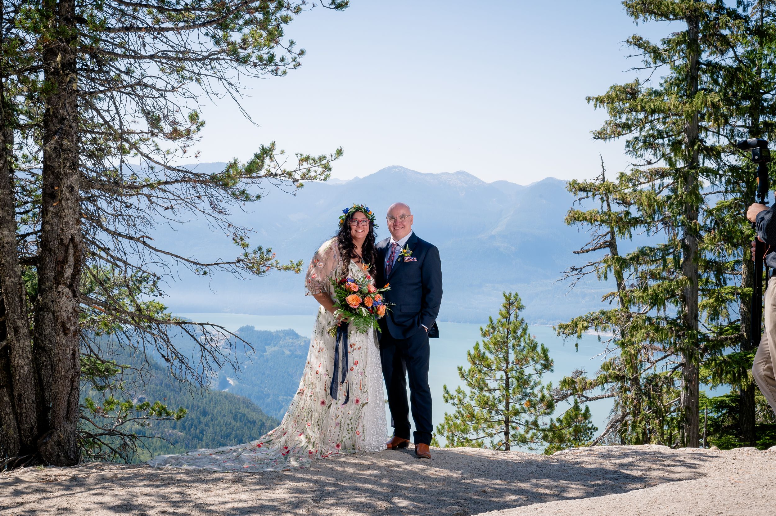 A couple in wedding attire stands on a mountain overlook with evergreen trees and snow-capped peaks in the background.