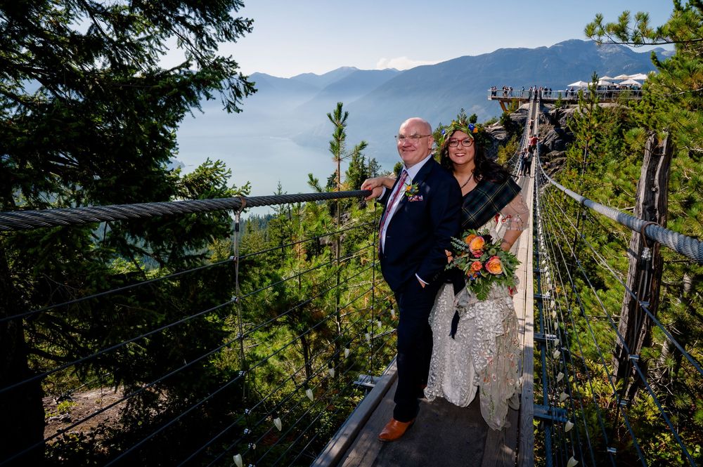 A couple shares a romantic moment on a scenic mountain suspension bridge surrounded by evergreen trees and distant peaks.