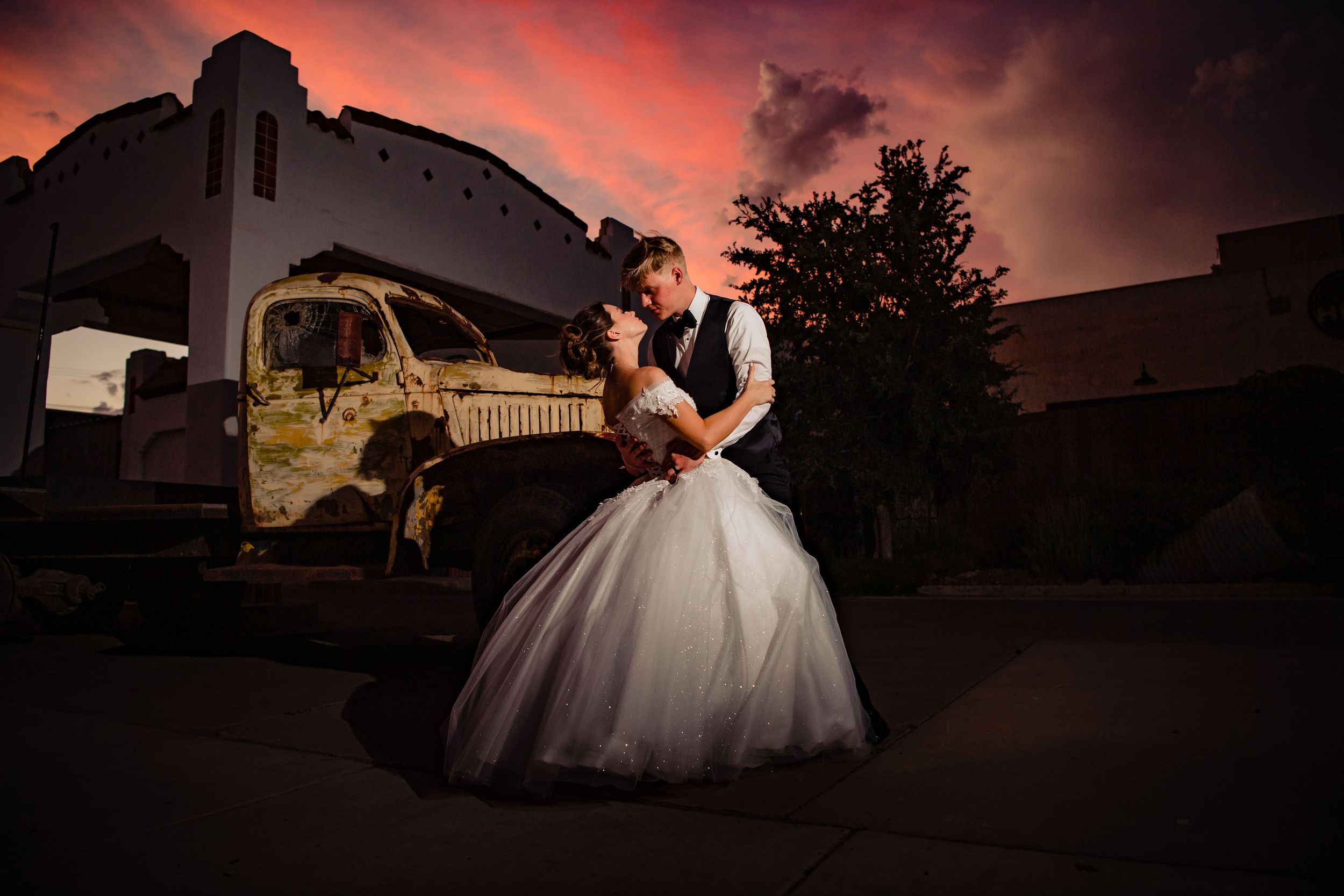 A couple embraces in front of an old train car during a dramatic sunset wedding photo shoot.
