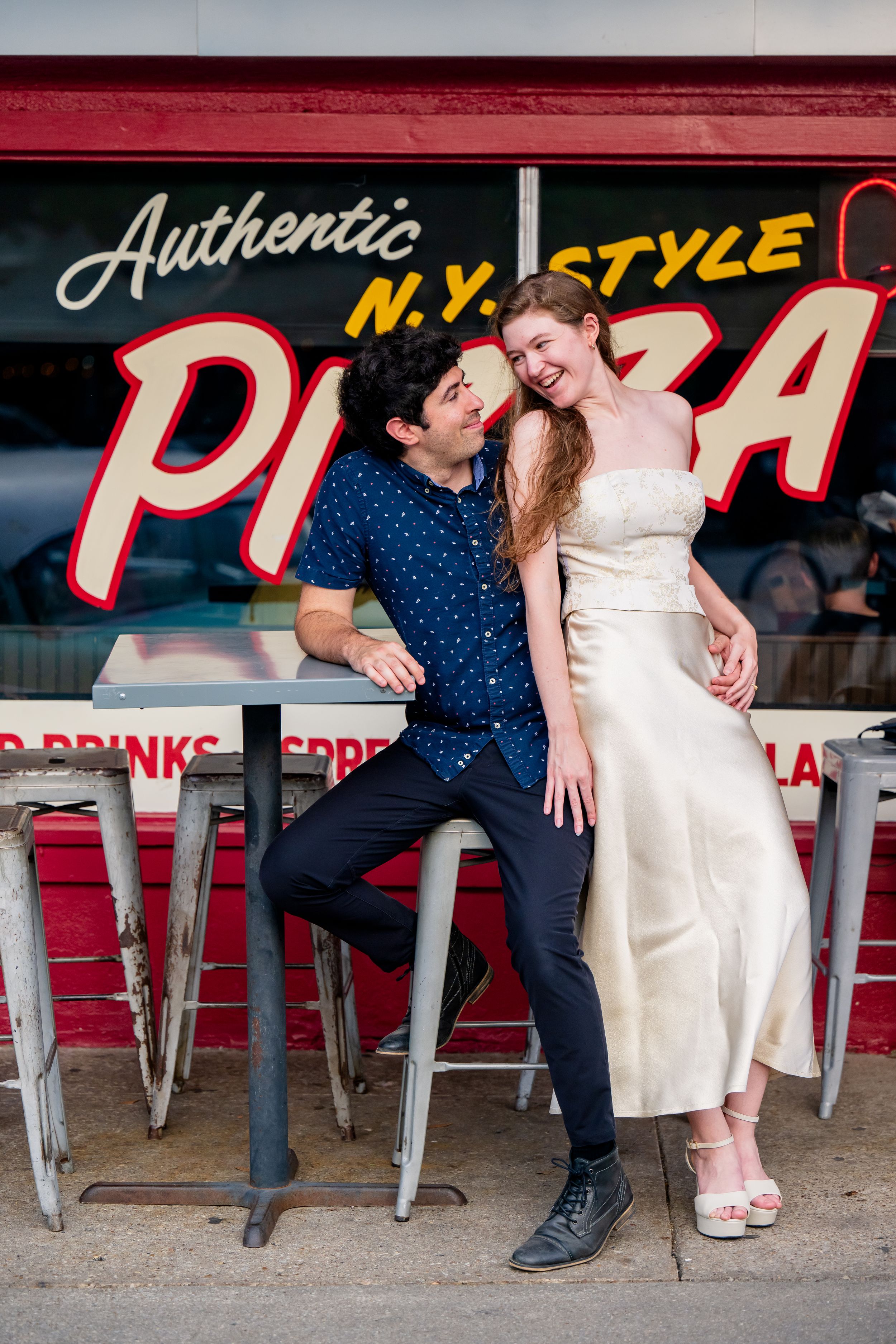 A couple poses together outside a New York style pizza restaurant with retro signage and bar stools.