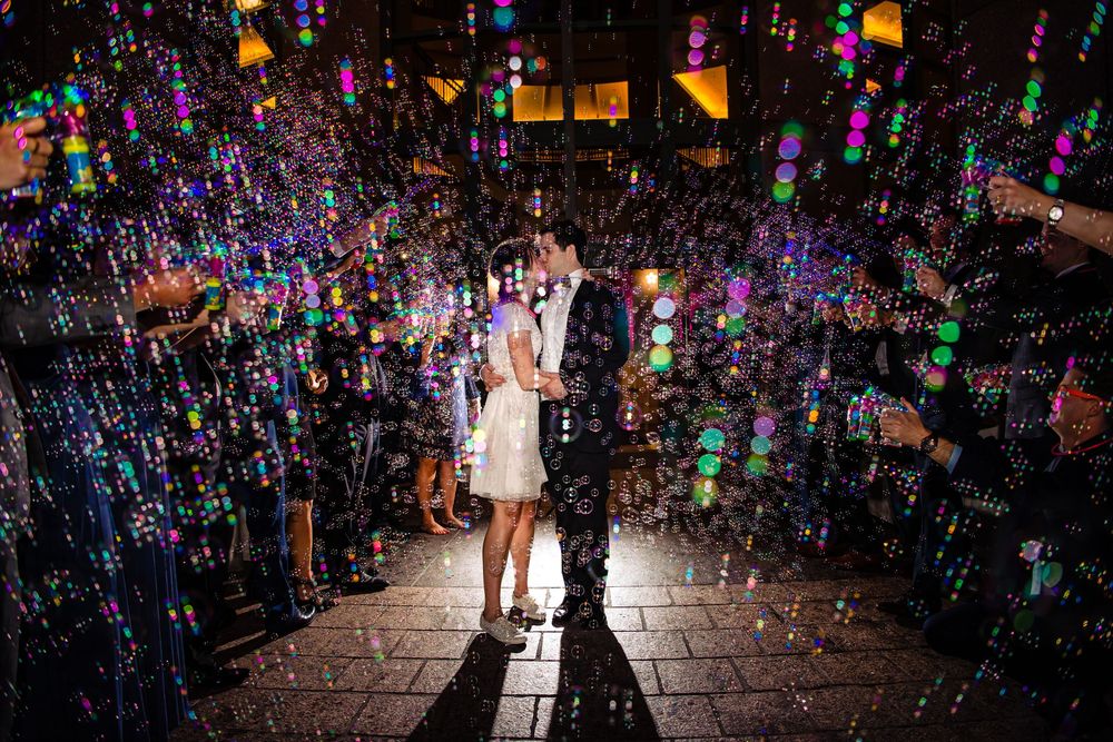 A couple stands embracing amid a magical display of colorful soap bubbles and lighting on a cobblestone street at night.