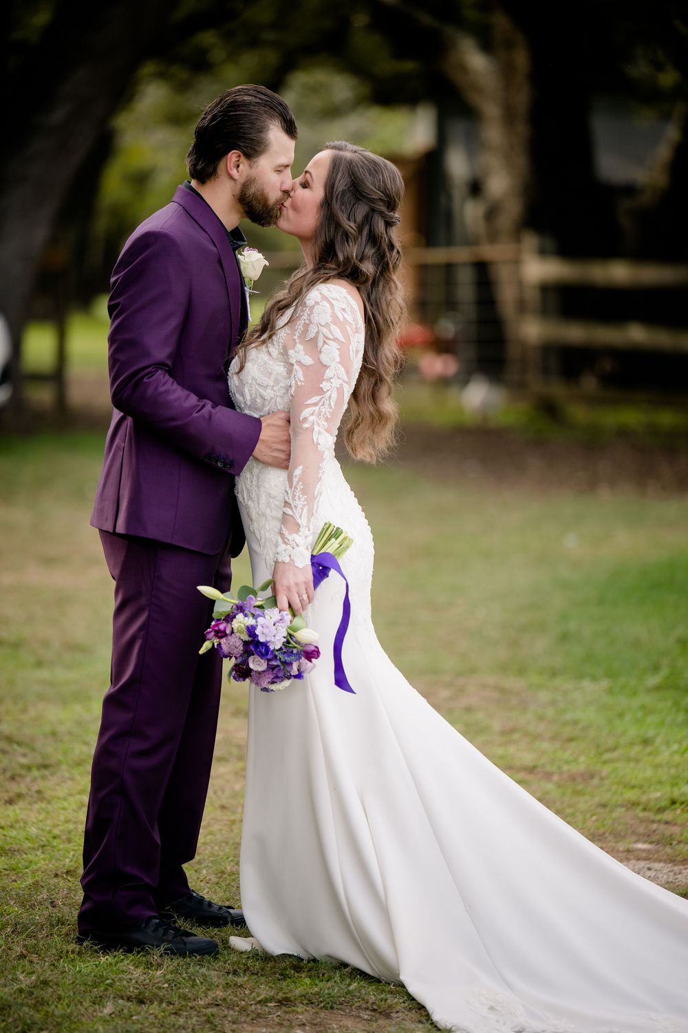 A couple in coordinated purple and white wedding attire share a romantic moment while holding a floral bouquet outdoors.