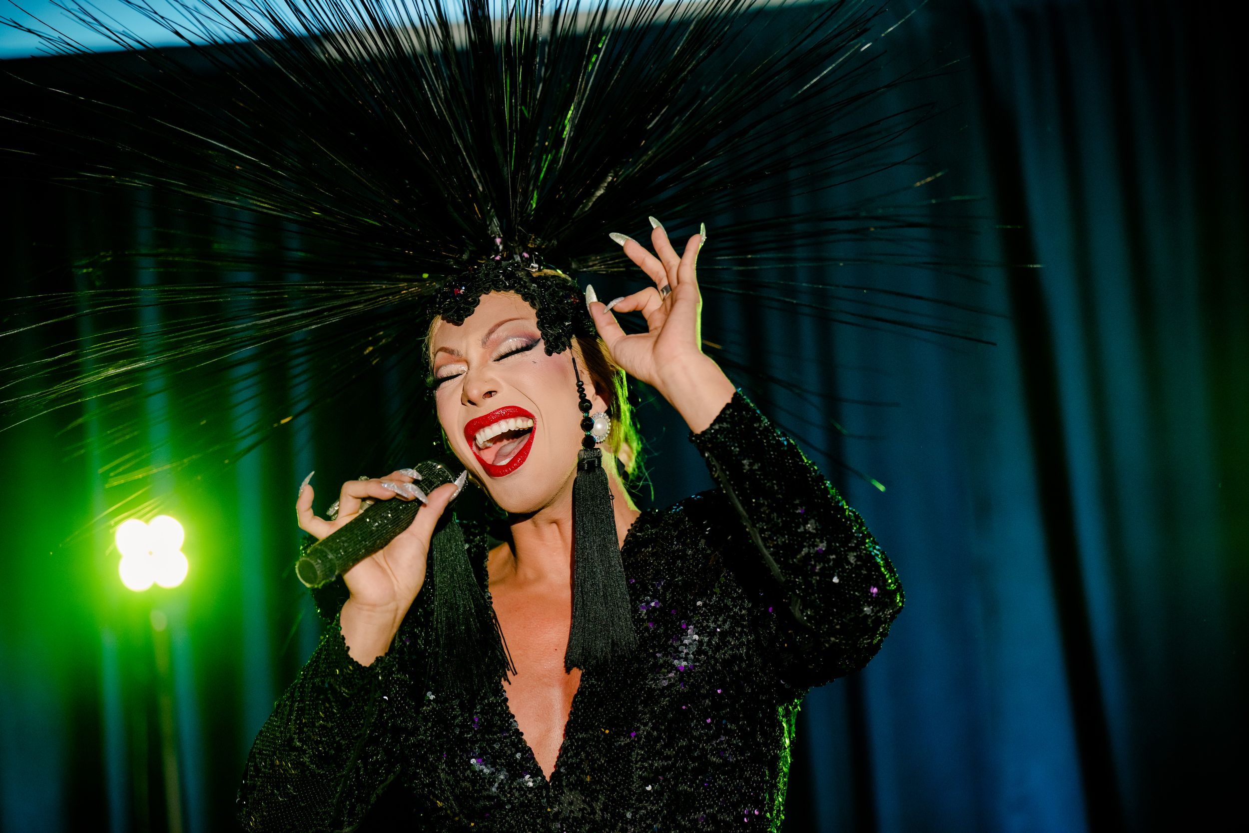 Performer in black sequin dress singing passionately on stage with bright green lighting and dark curtain backdrop.