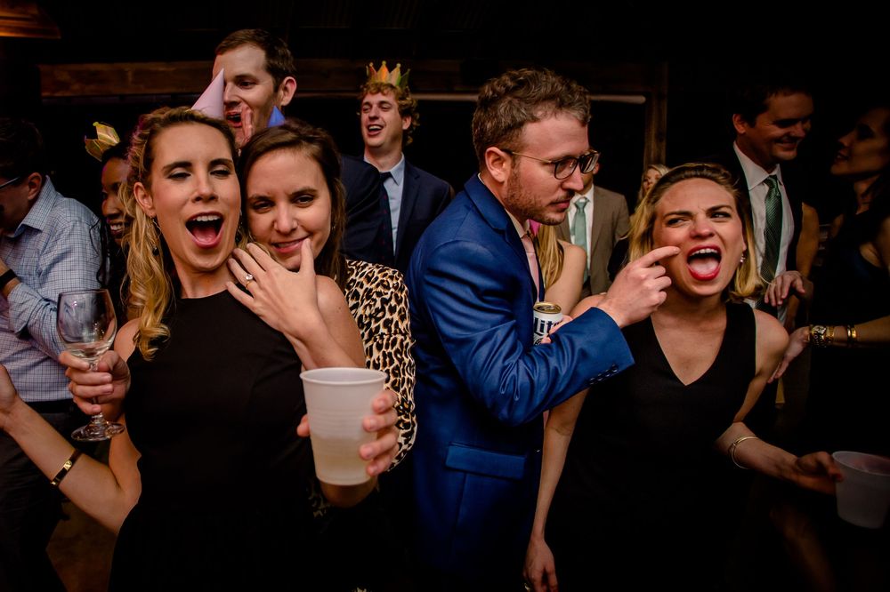 Party guests show energetic expressions while dancing and celebrating at a dimly lit indoor reception event.