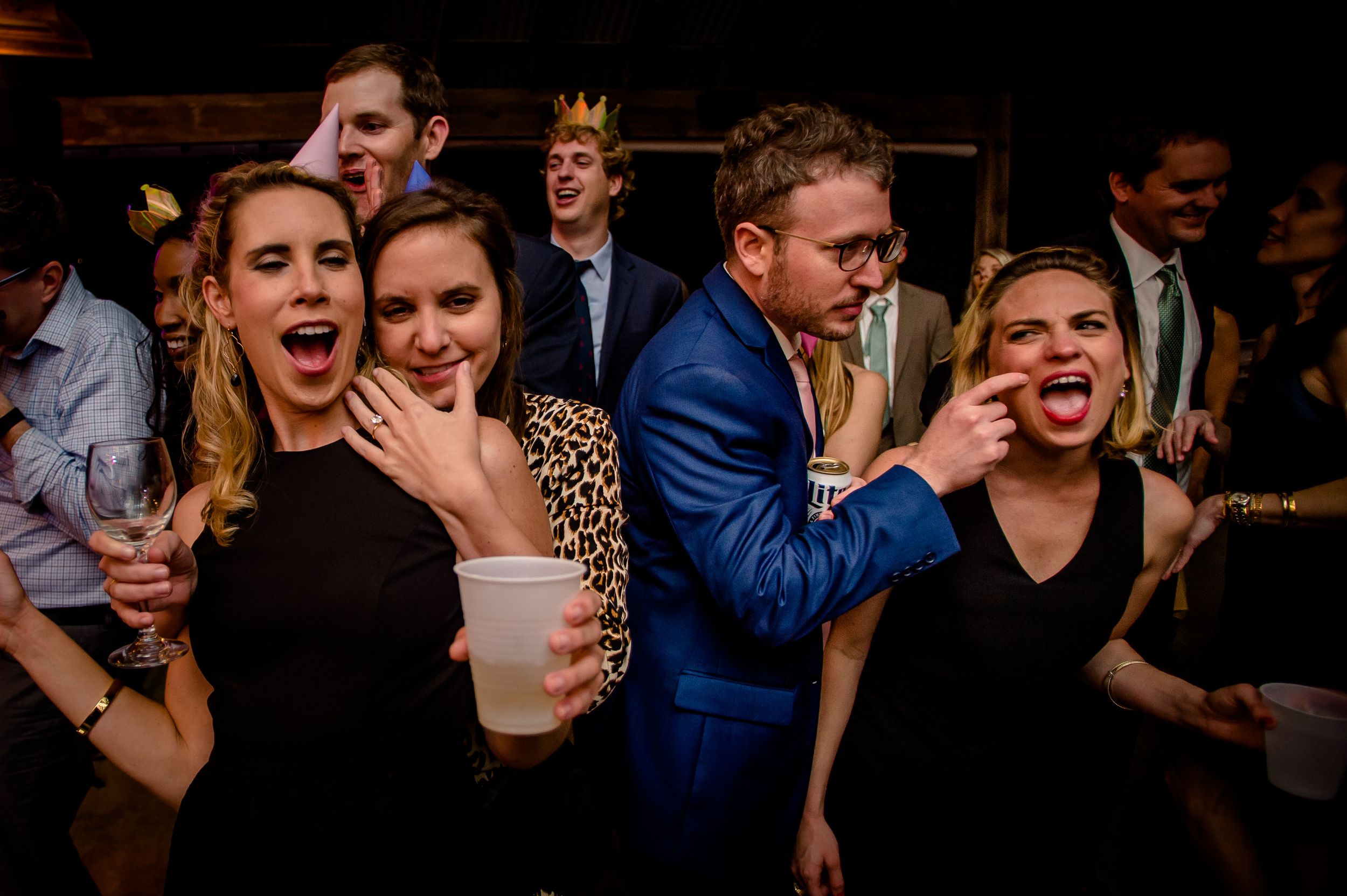 Party guests show energetic expressions while dancing and celebrating at a dimly lit indoor reception event.