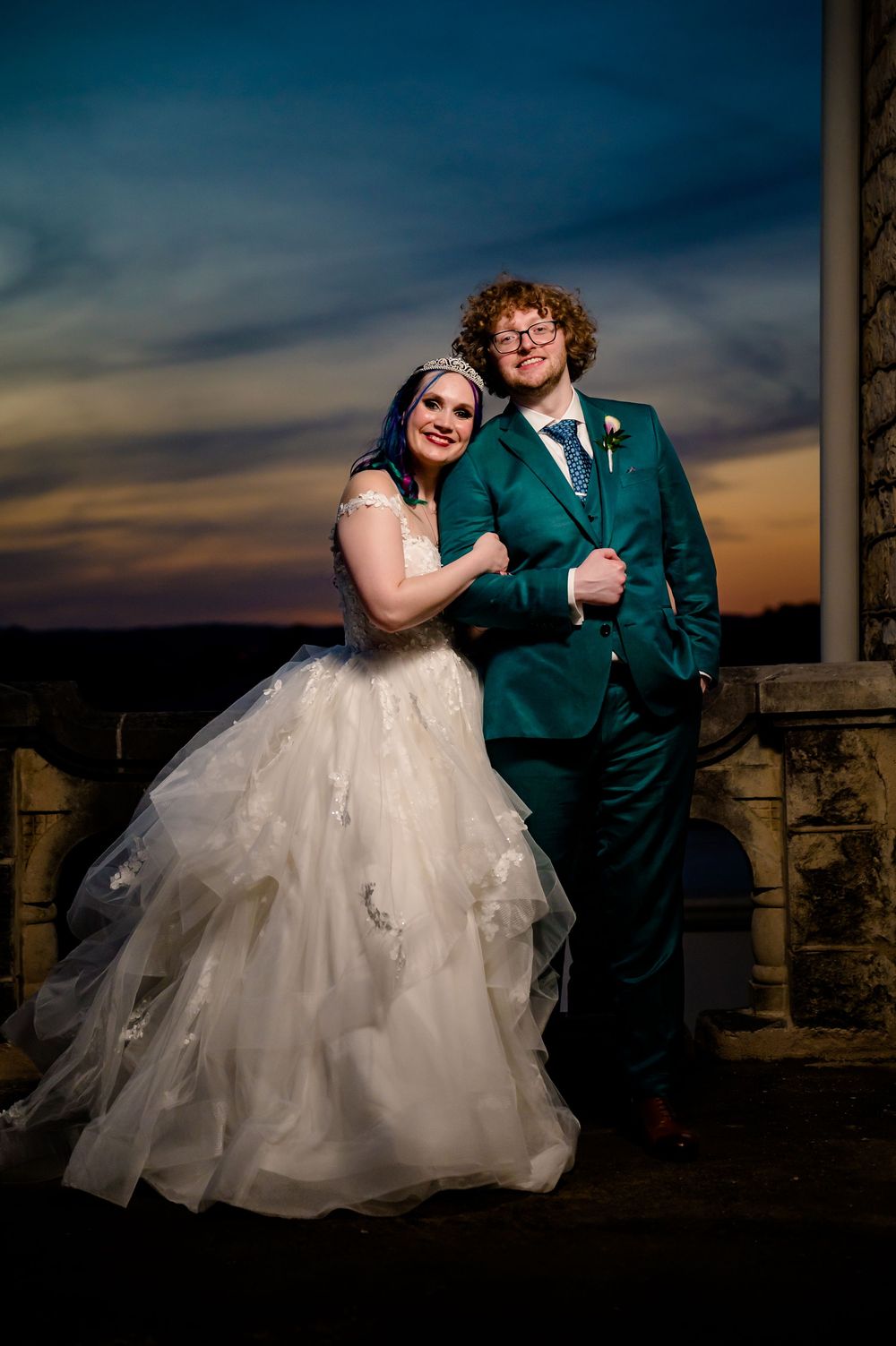 A wedding couple poses at sunset against stone architecture, with the bride in white and groom in emerald green suit.
