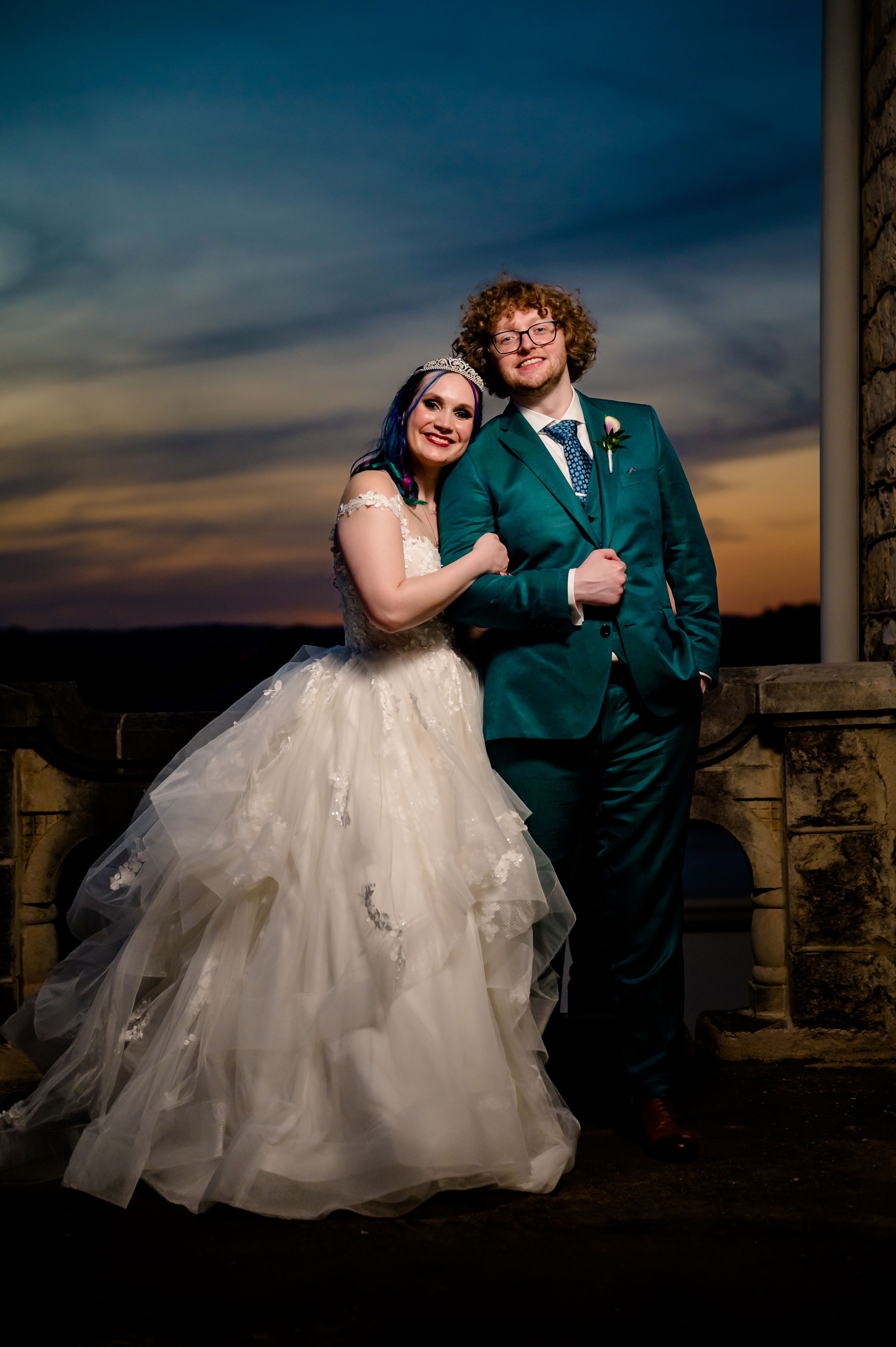 A wedding couple poses at sunset against stone architecture, with the bride in white and groom in emerald green suit.