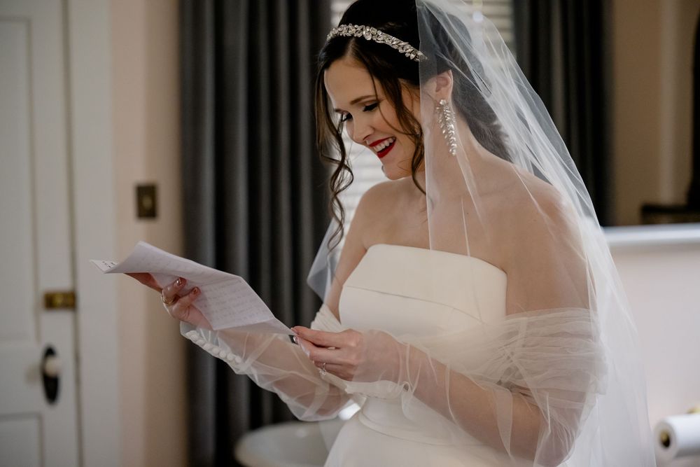Bride in a strapless white wedding gown and veil reads a letter while getting ready for her ceremony.