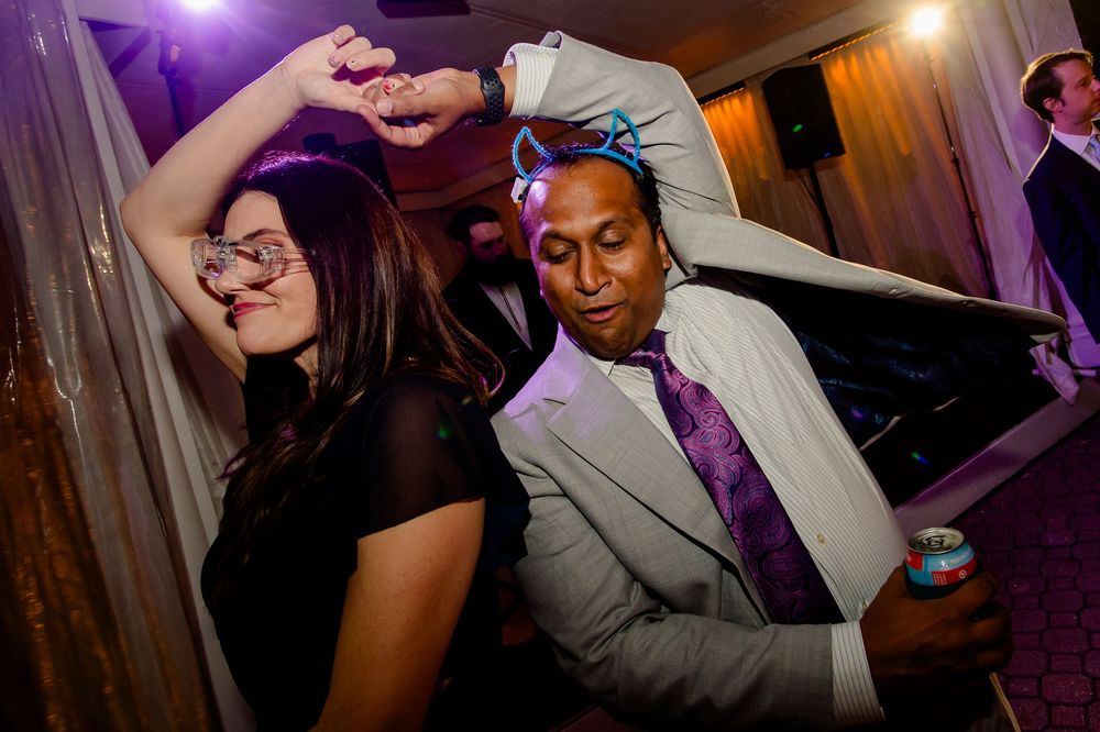 Party guests dance enthusiastically at a celebration with purple lighting and festive atmosphere in a wood-paneled room.