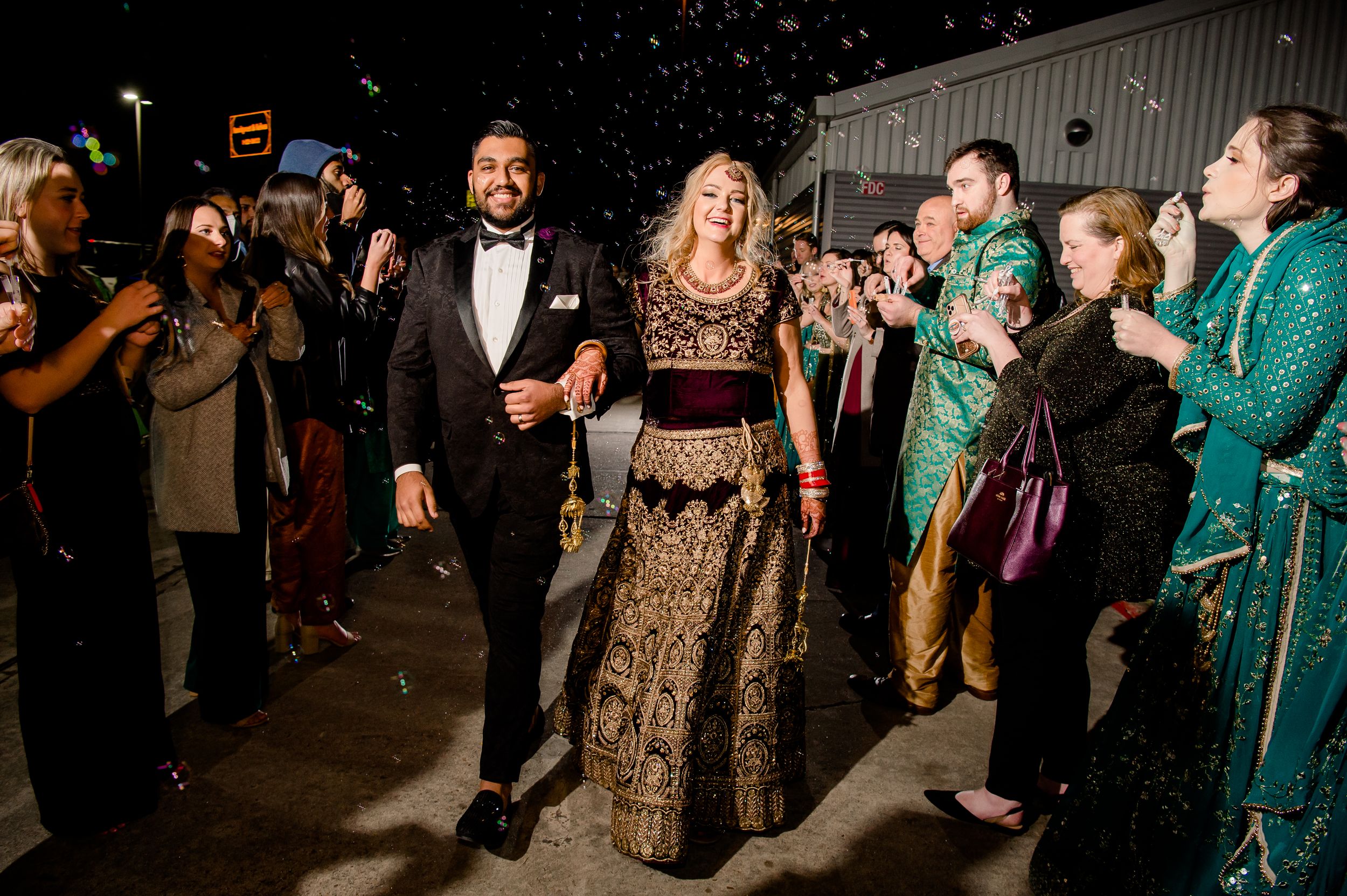 A couple in formal black and gold attire walk through sparklers at night while celebratory guests surround them.