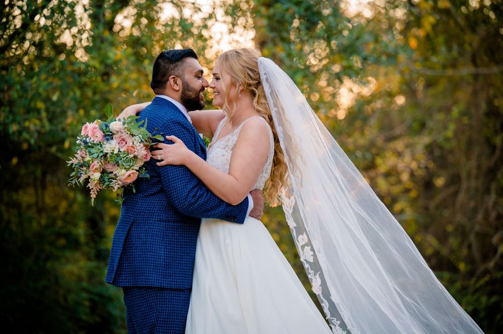 Two figures in wedding attire embrace in a romantic moment at sunset with a flowing white veil.