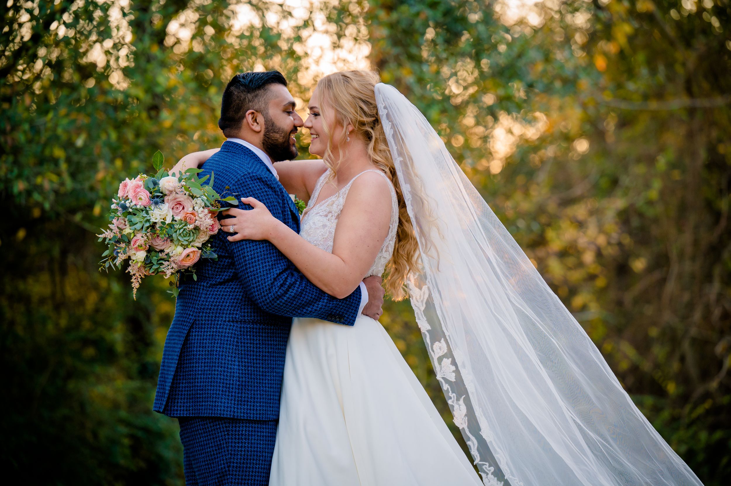 Two figures in wedding attire embrace in a romantic moment at sunset with a flowing white veil.