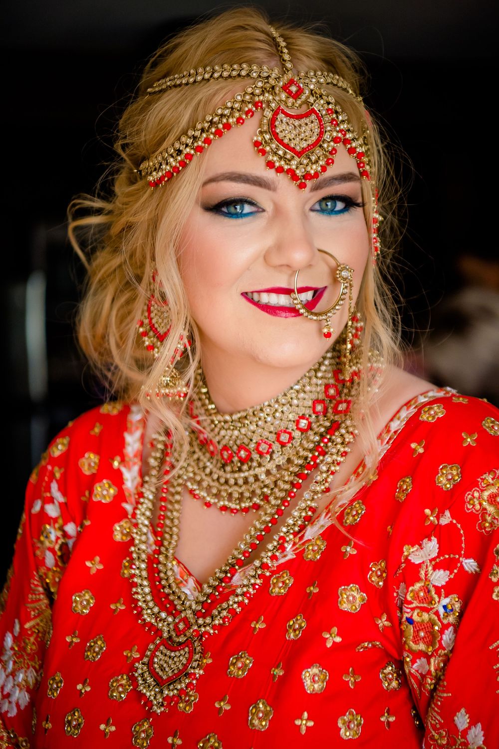 Bride wearing a traditional red and gold Indian wedding ensemble with ornate jewelry and headpiece at a celebration.