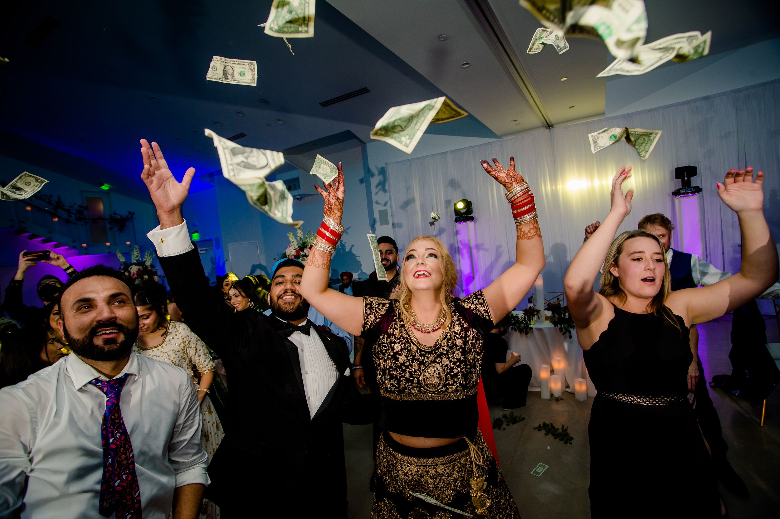 Wedding guests dancing and throwing money in the air while celebrating on a dimly lit dance floor during a reception.