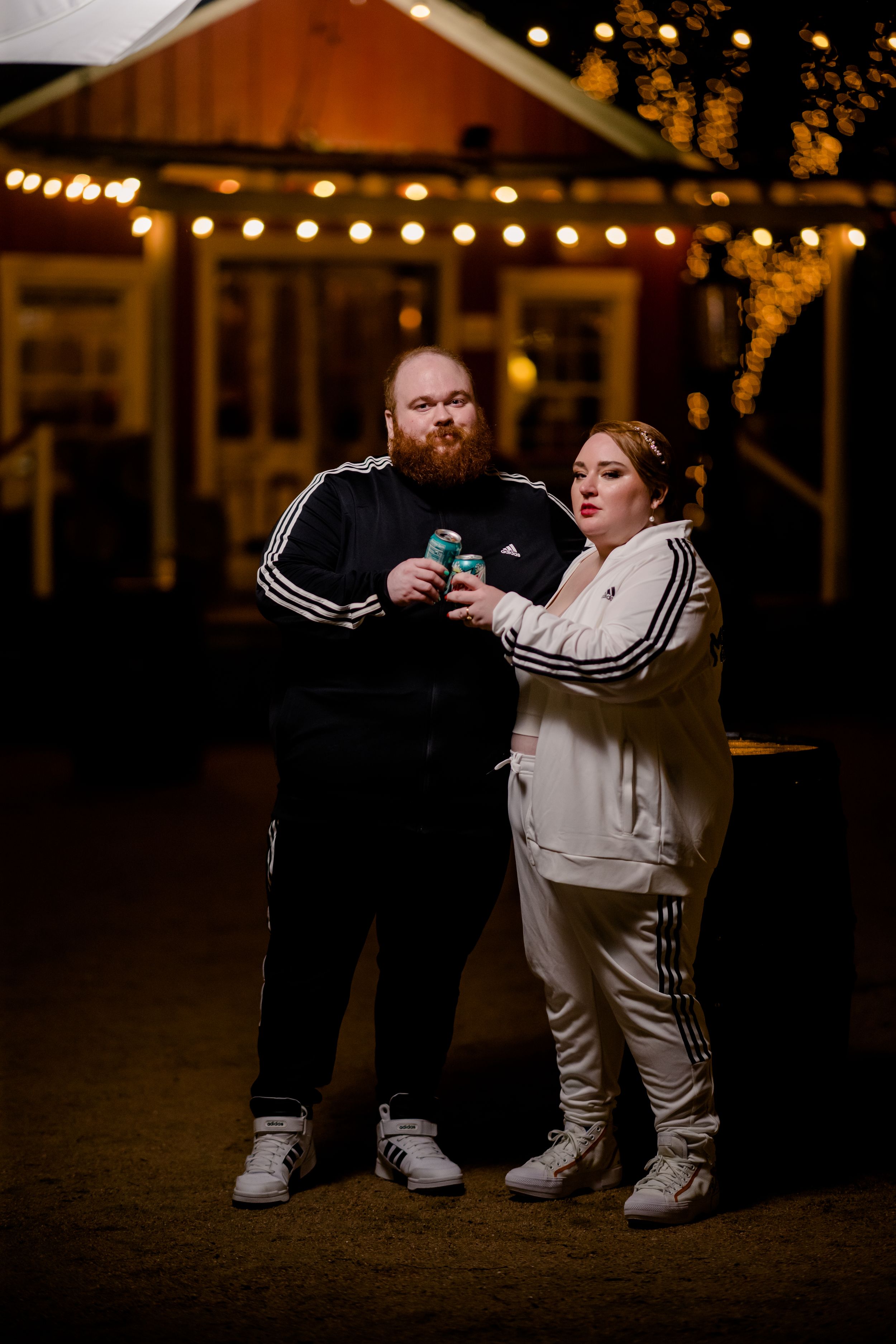 A couple wearing matching Adidas tracksuits pose together at night with twinkle lights in the background.