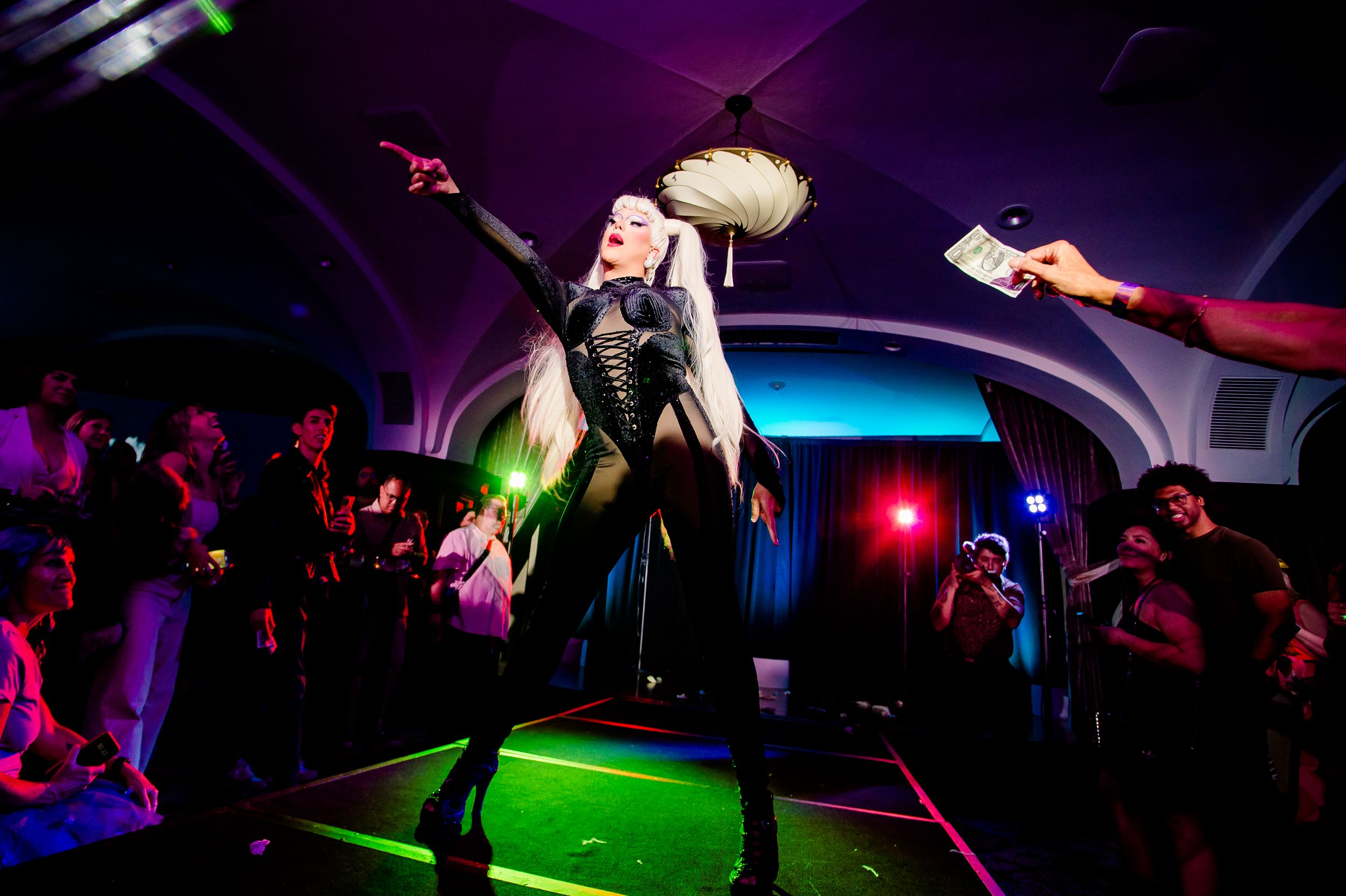 A performer in black and blonde hair strikes a dramatic pose on a lit-up green platform with a purple-lit crowd in background.