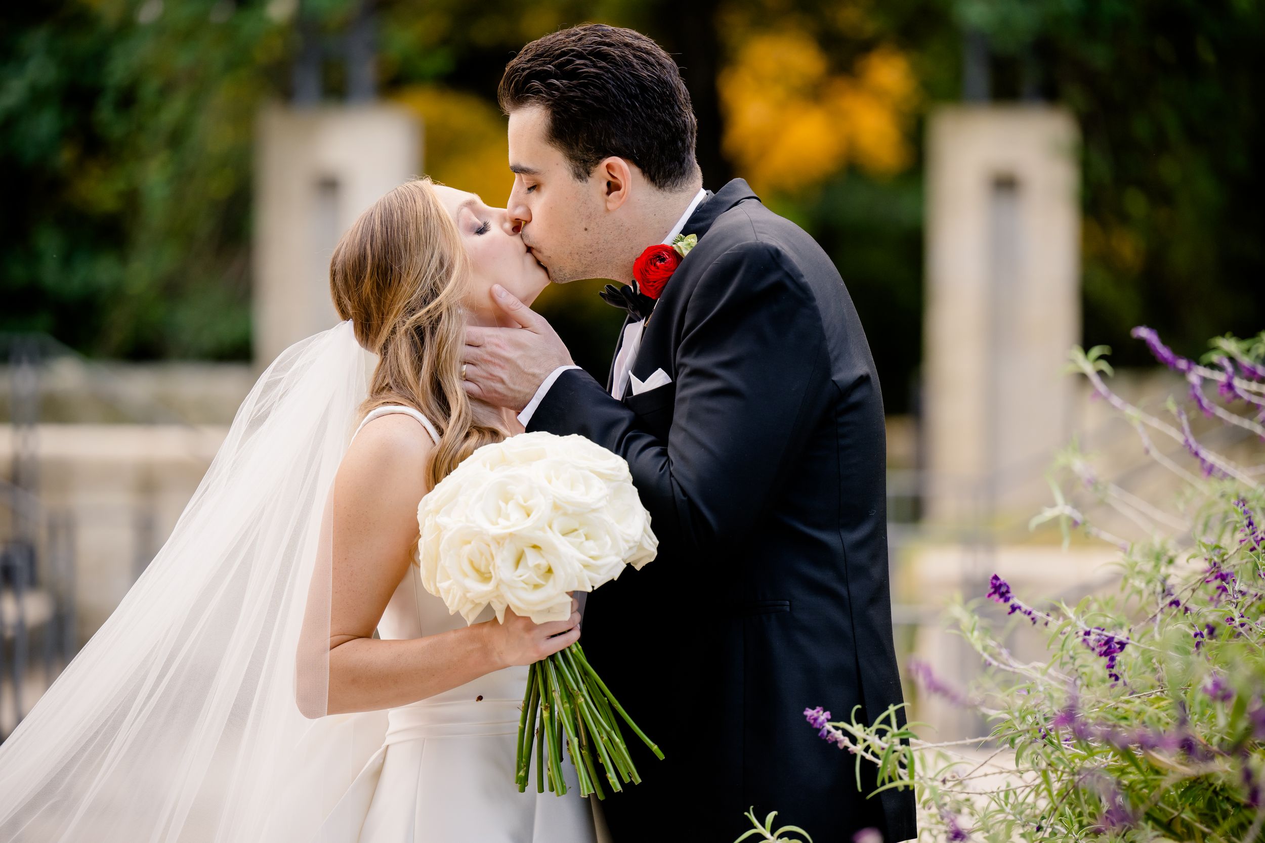 A romantic wedding couple shares a kiss while holding a white bouquet in a garden with blooming flowers.