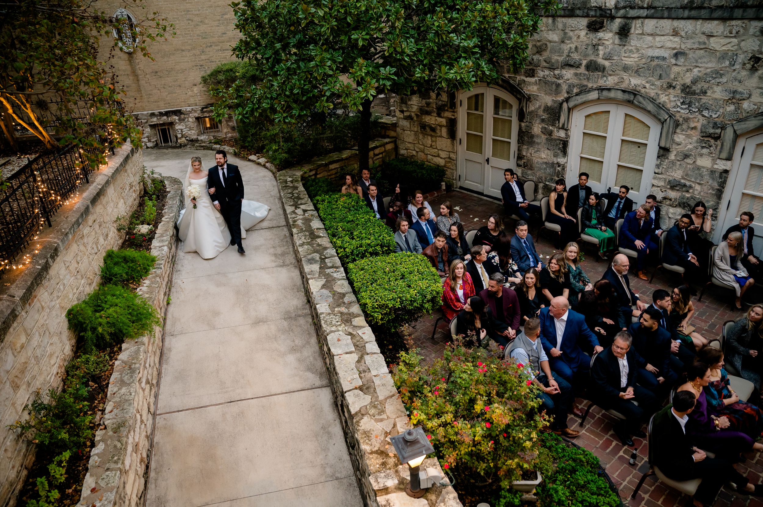 A couple walks through a garden path while wedding guests watch from below at an elegant stone venue ceremony.