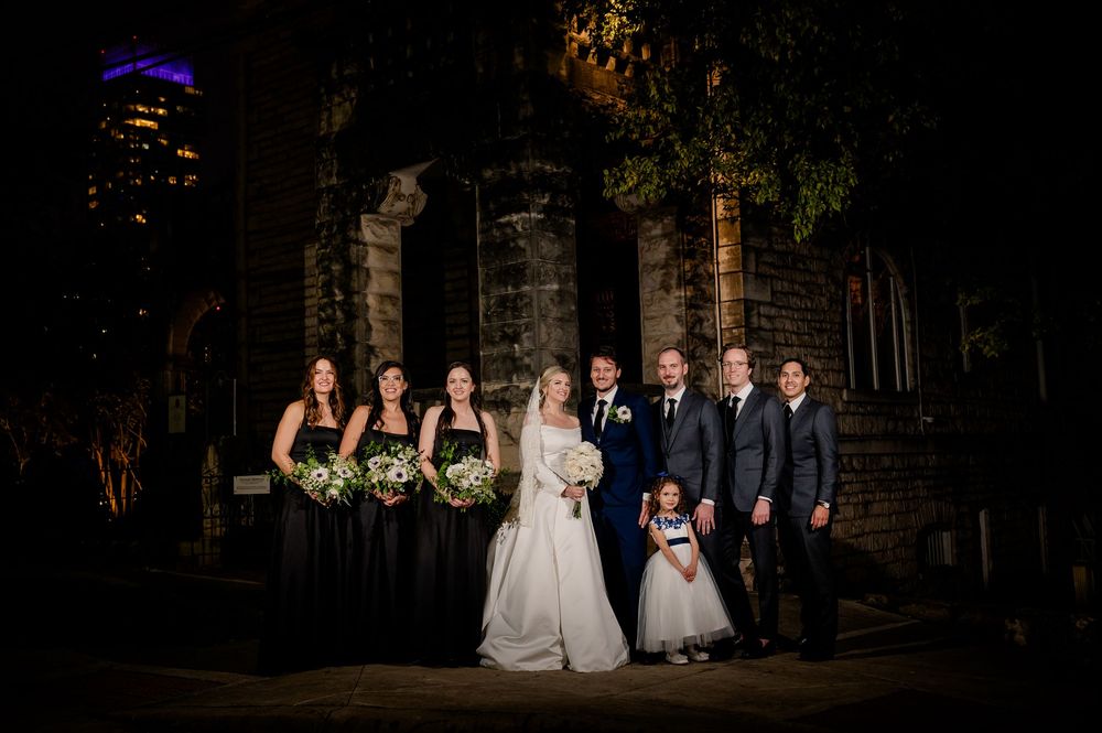 Wedding party poses at night outside stone church with city lights glowing in background.
