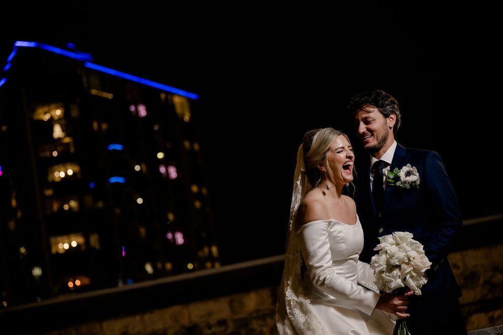 A bride in a strapless wedding dress laughs joyfully while holding a white bouquet during an outdoor nighttime celebration.