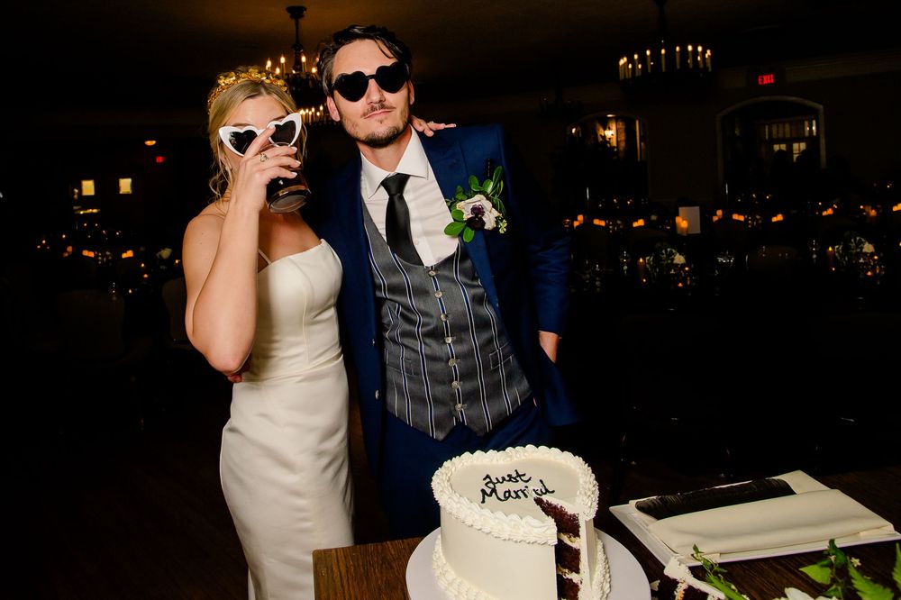 A newlywed couple in sunglasses poses playfully next to their wedding cake at an evening reception.