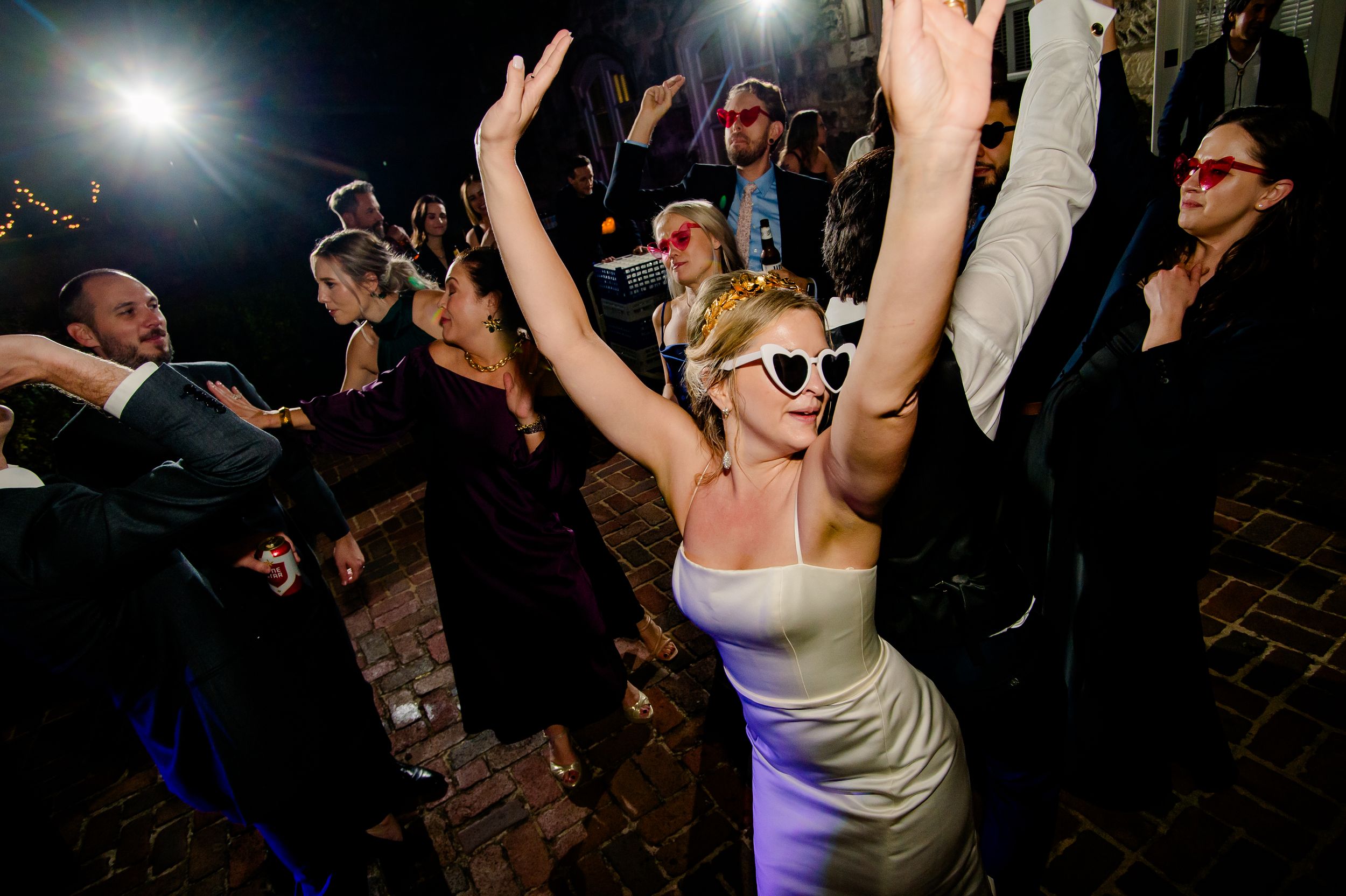 Enthusiastic partygoer wearing sunglasses and a purple dress dances with raised arms on a crowded dance floor.