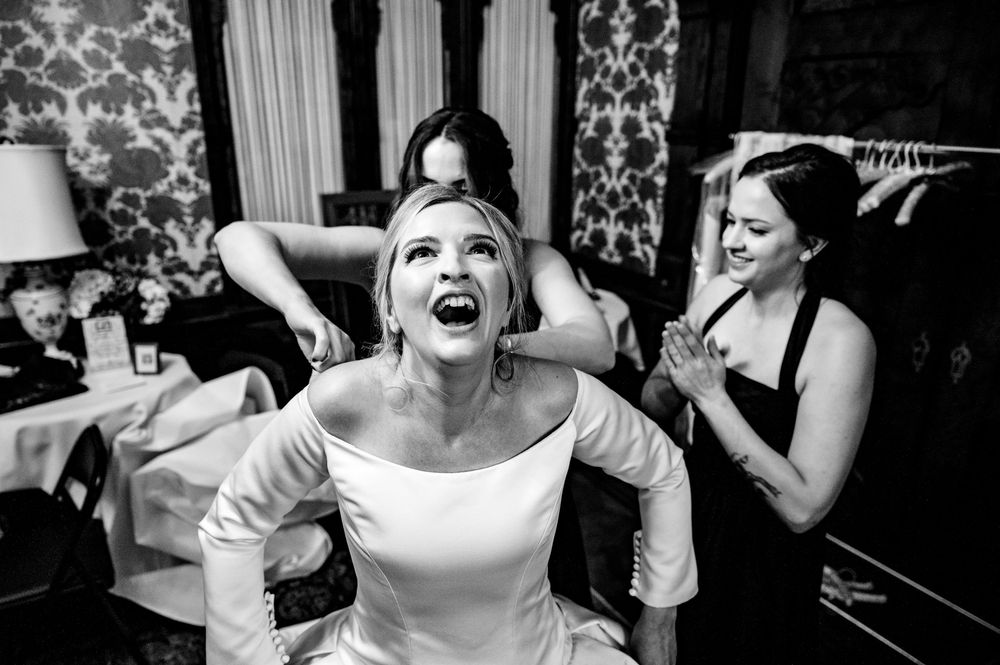 Black and white candid photo showing a joyful moment during wedding preparations in an ornate room with patterned wallpaper.