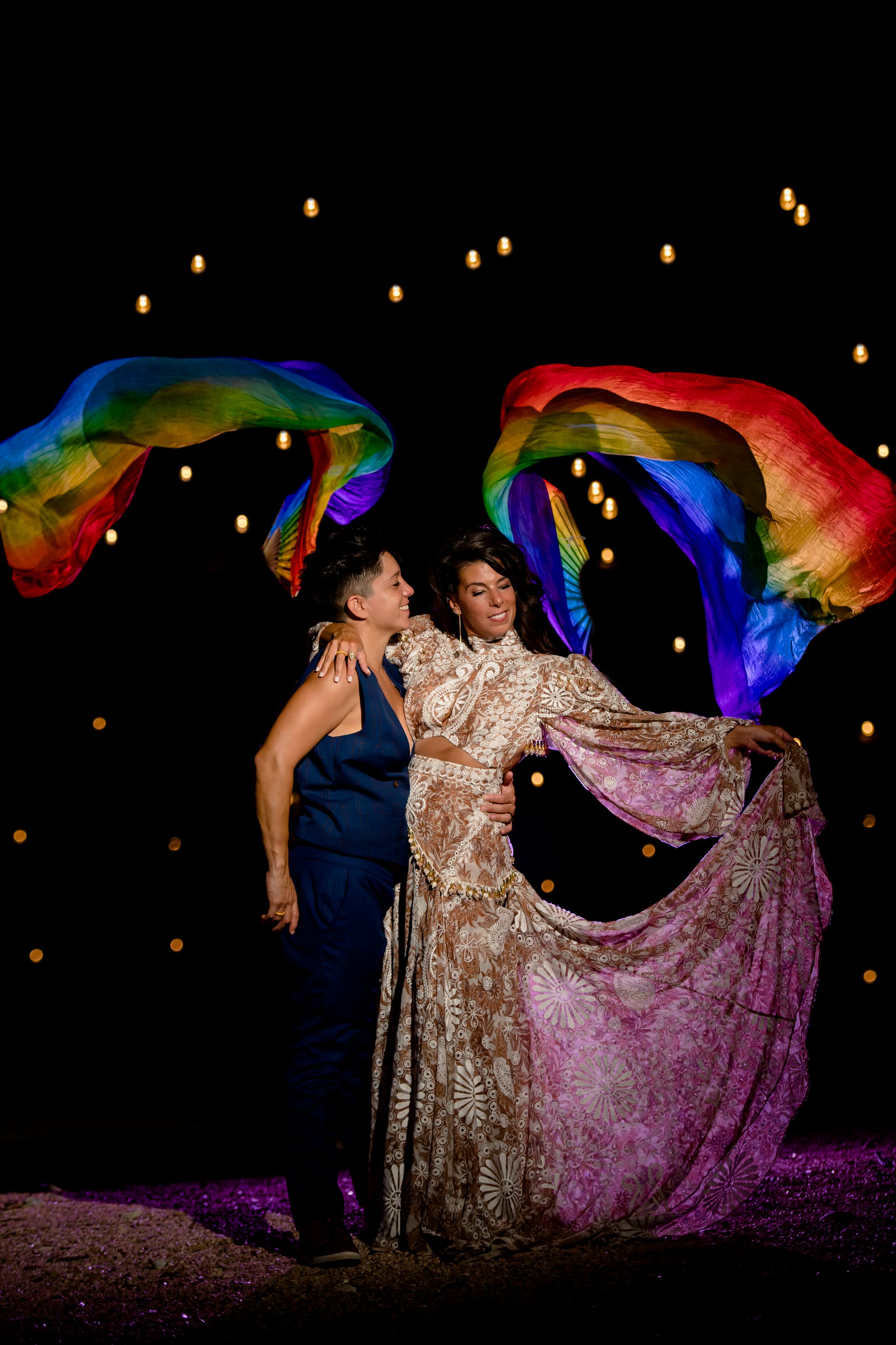 Two people dance together at night under glowing rainbow fabric banners with floating lanterns in the background.