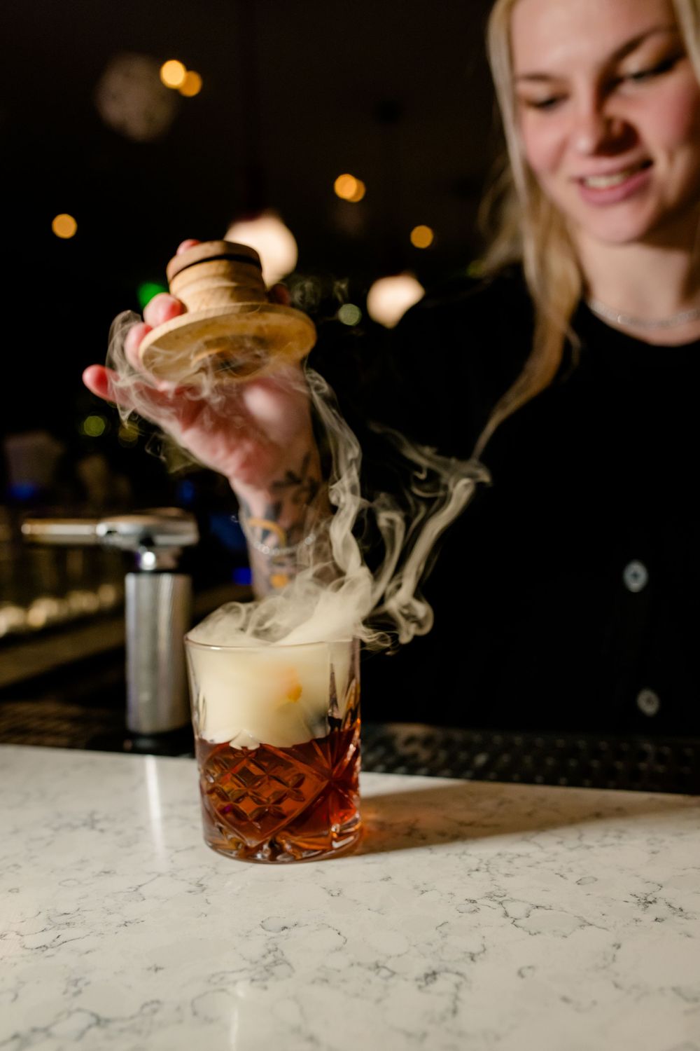 A dramatic cocktail presentation shows smoke being captured under a glass dome on a marble bar counter.