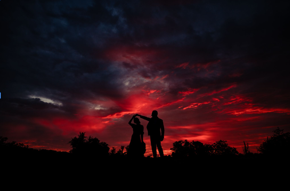 Silhouettes of people stand against a dramatic red and black sunset sky with clouds.