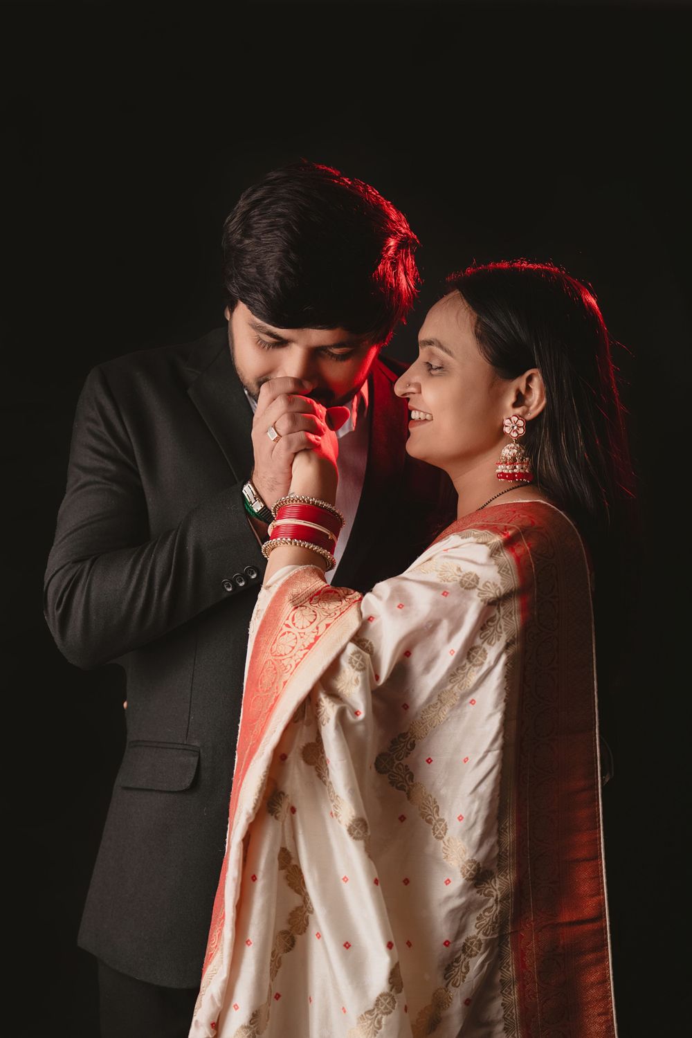 A romantic couple poses against dark background wearing traditional Indian attire with red accents.