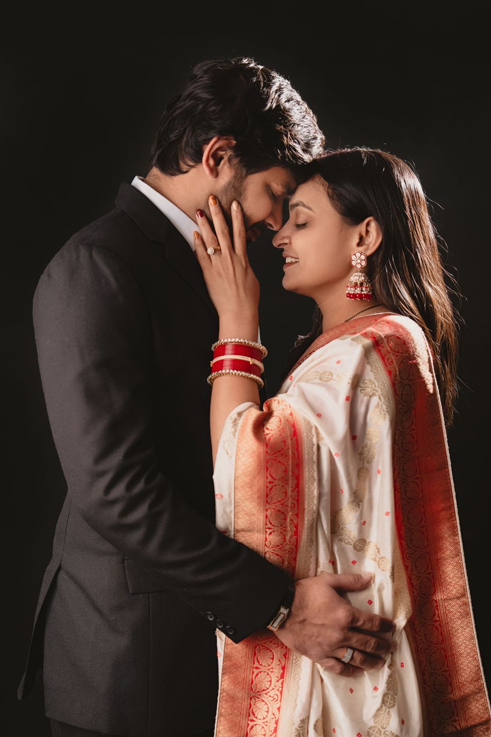 An intimate portrait of a couple in formal Indian wedding attire sharing a tender moment against black background.