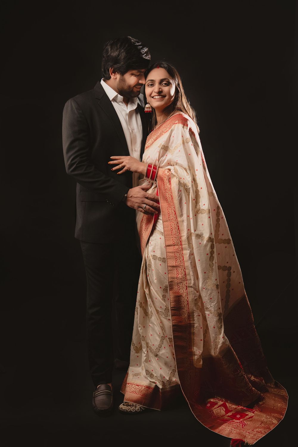 A stylish couple in contrasting black suit and cream saree pose dramatically against dark backdrop.