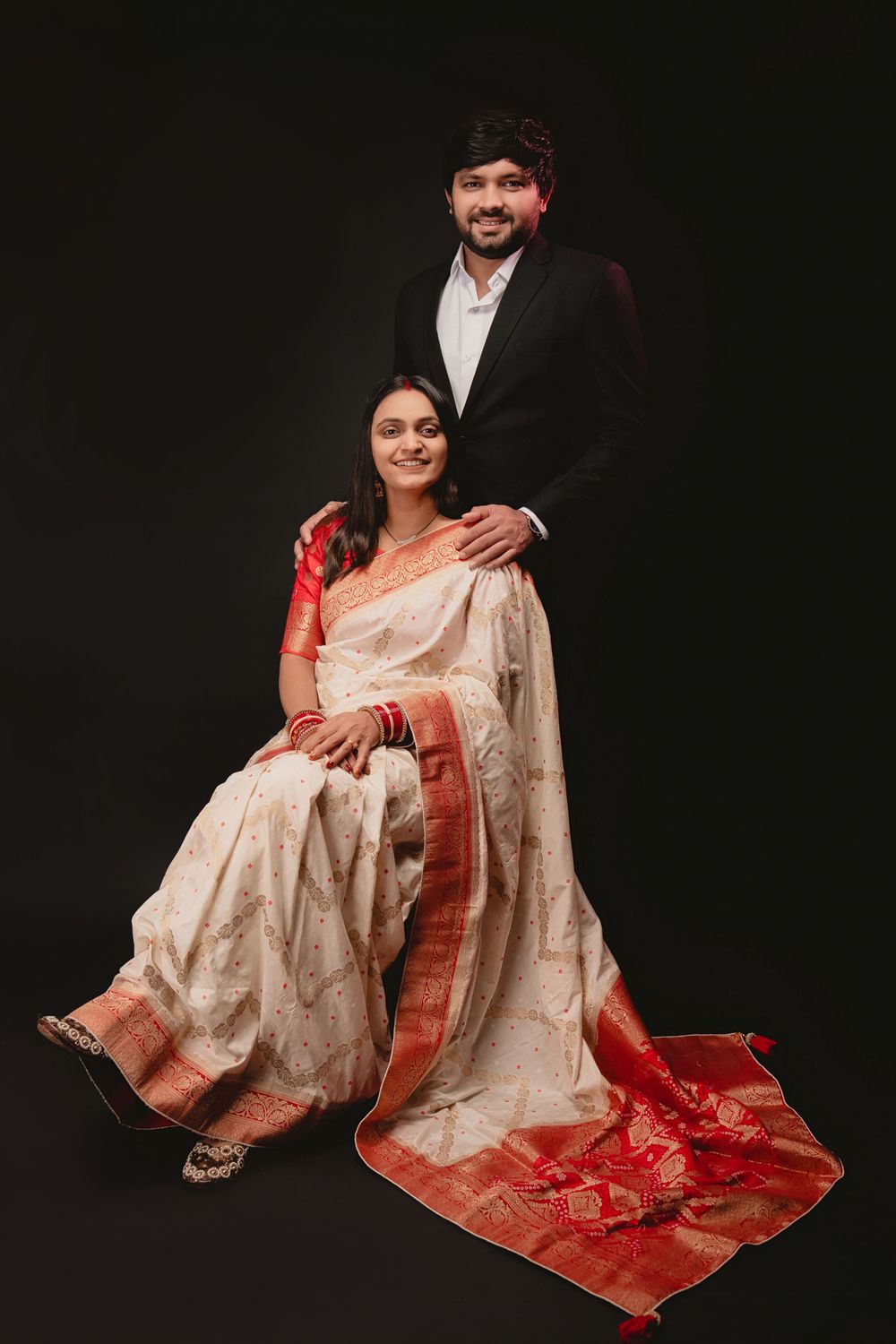 Couple wearing traditional Indian attire poses elegantly against dark backdrop.