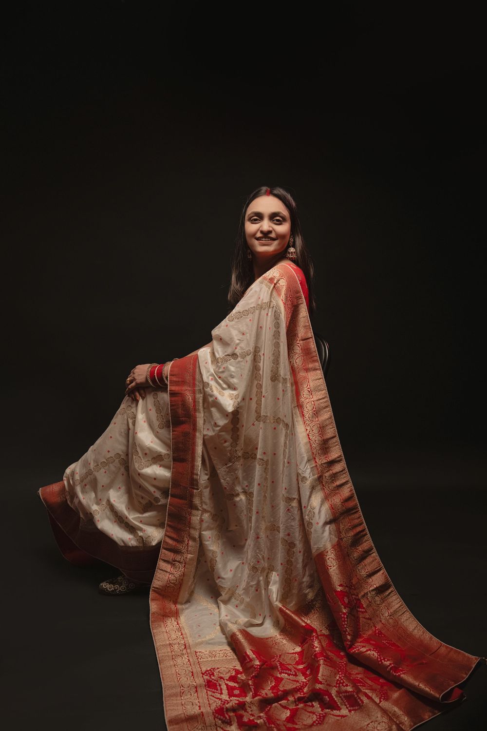 Woman in elegant cream and red silk saree poses gracefully against dark backdrop during traditional photoshoot.