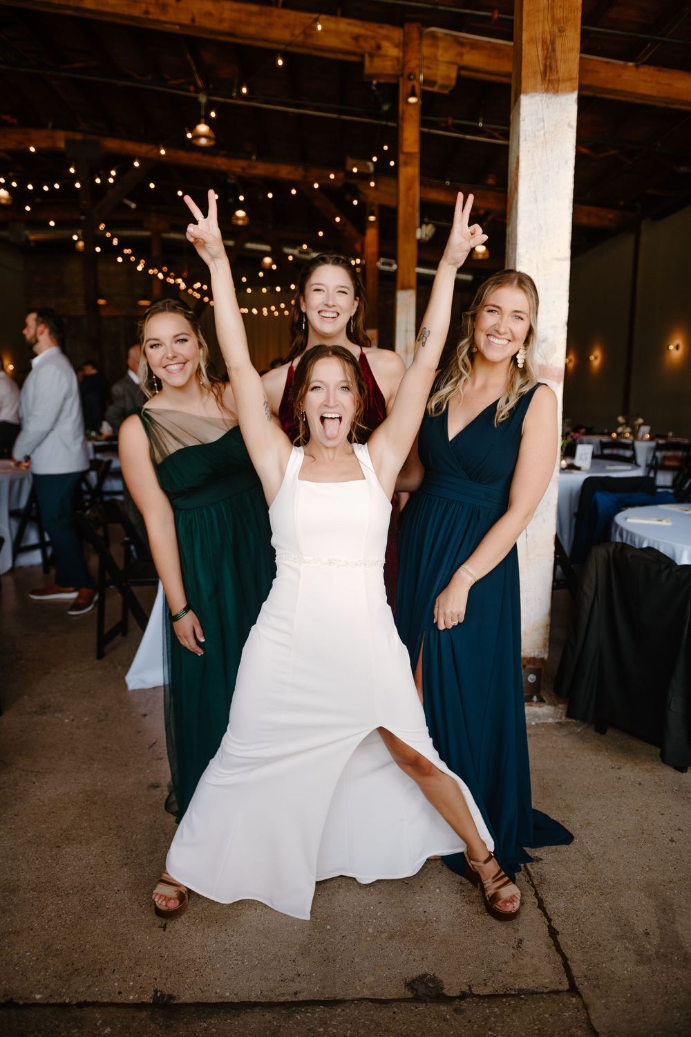 Wedding party members in teal dresses celebrate joyfully with the bride at an indoor reception venue with string lights.