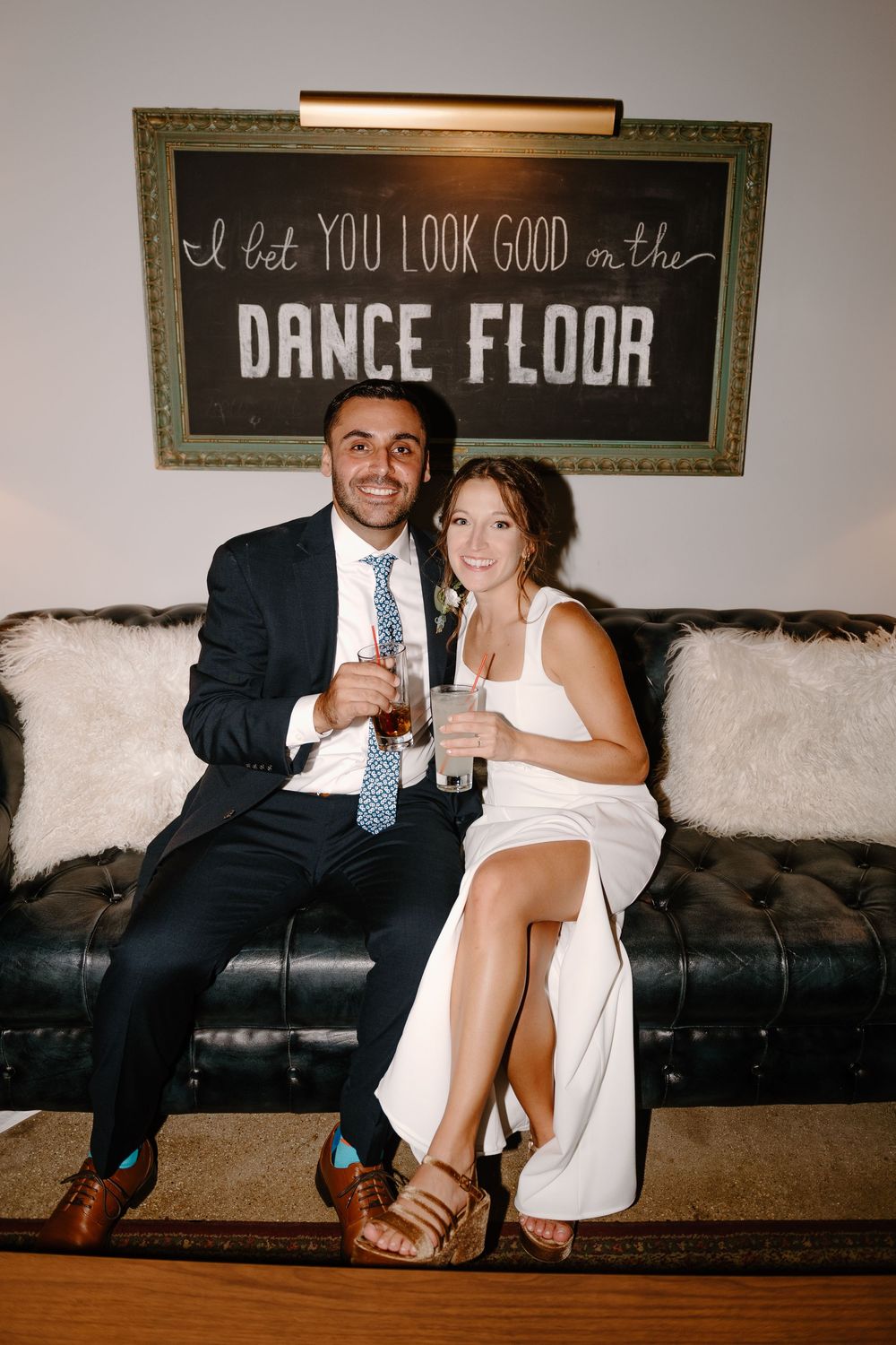 Couple sits intimately on dark leather couch under rustic wall signs at wedding reception venue.