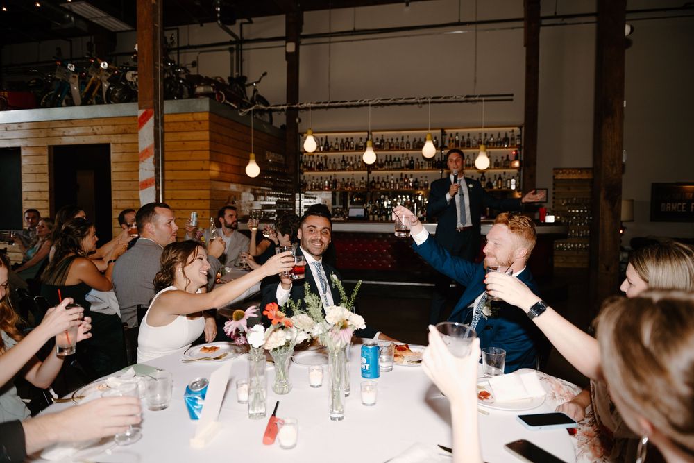 Wedding guests raise their glasses in a toast at a reception dinner in an industrial-style venue with hanging lights.